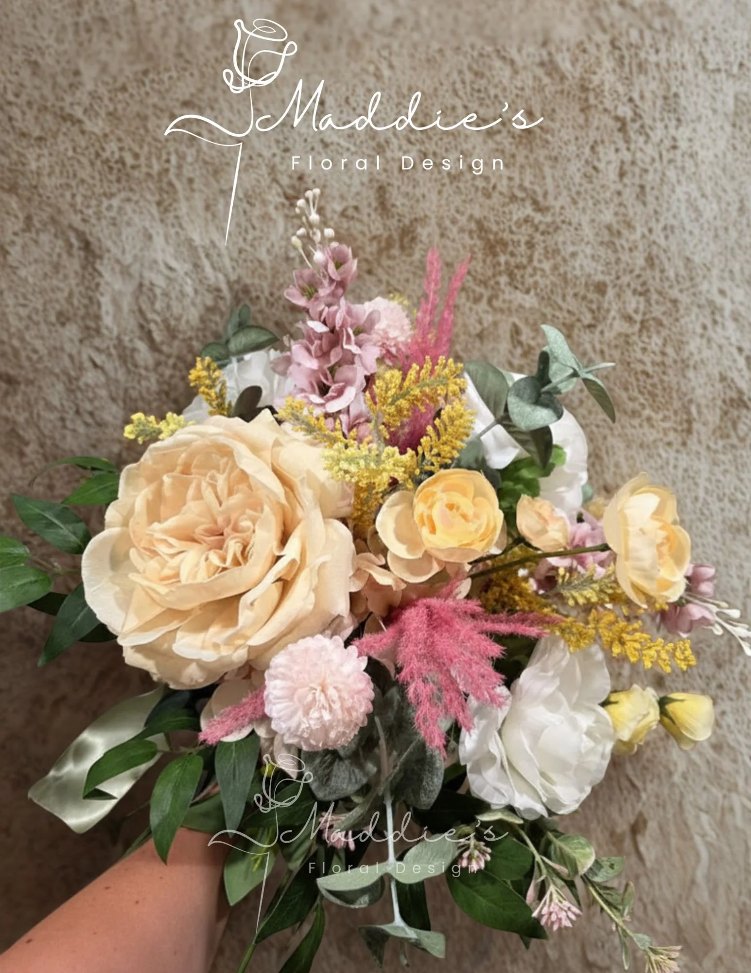 A bouquet of various pink, peach, white, and yellow flowers with green leaves, held in a person's hand against a textured beige wall. The image has text overlay indicating "Maddie's Floral Design."