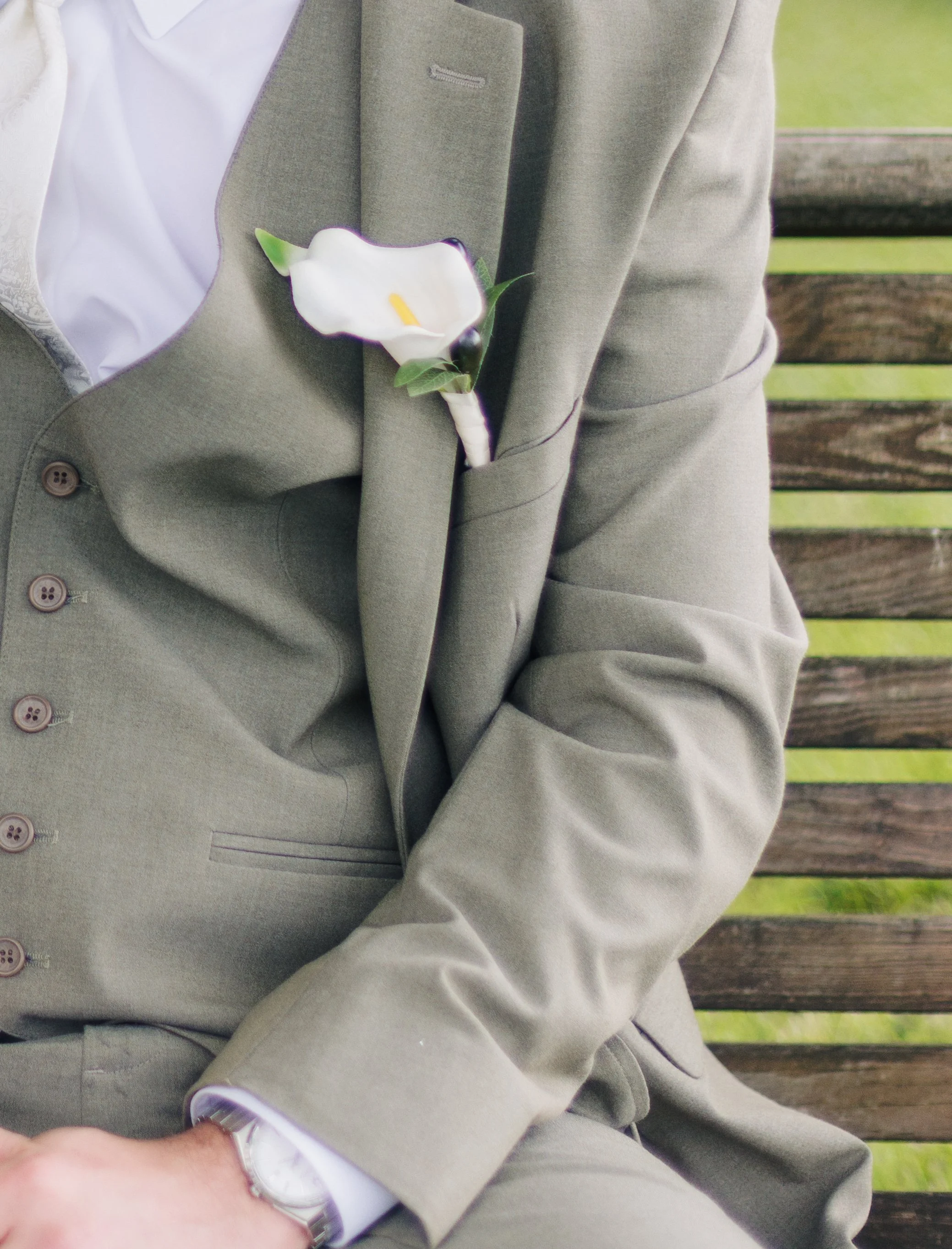 Close-up of a man's torso wearing a gray suit with a white shirt and a boutonnière flower in the lapel pocket, sitting on a wooden bench with green grass in the background.