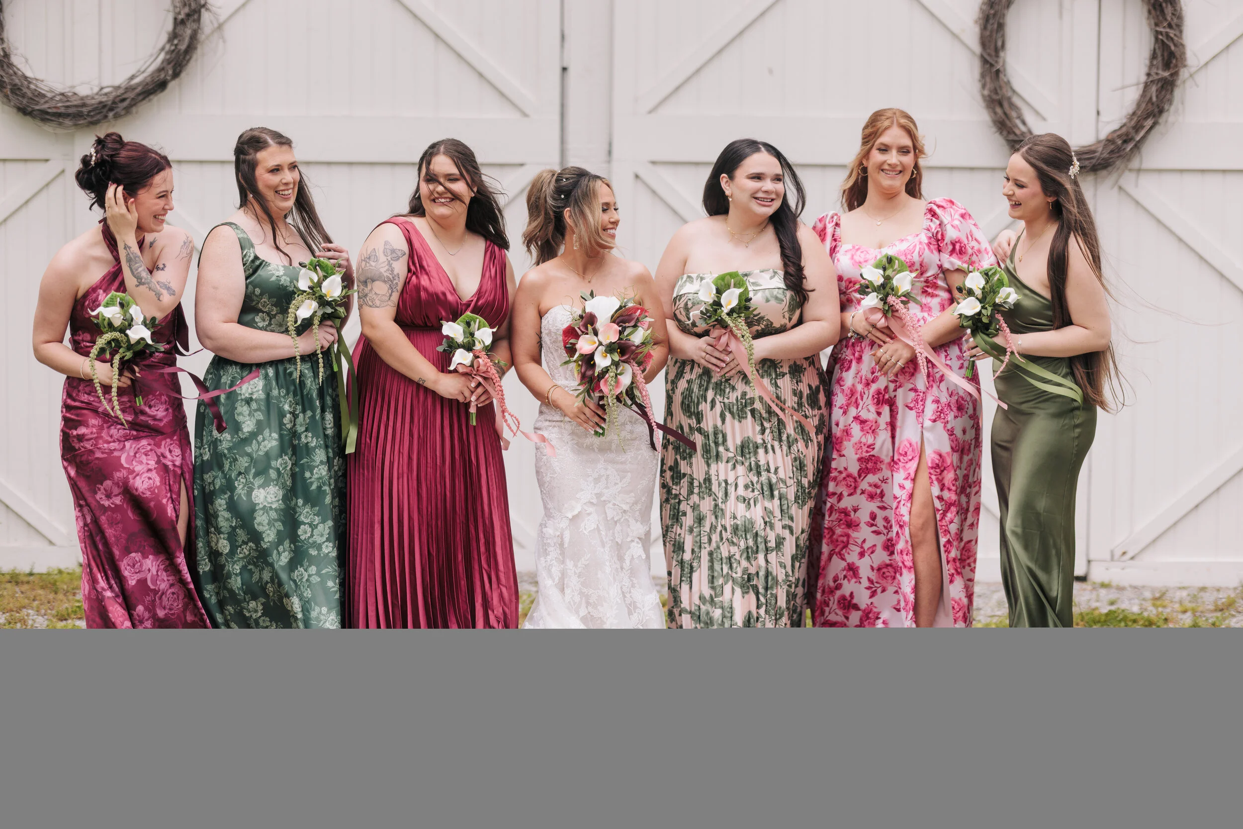 A group of eight women in colorful dresses holding bouquets of flowers, standing in front of white barn doors with wreath decorations, smiling and laughing together at a wedding or celebration.