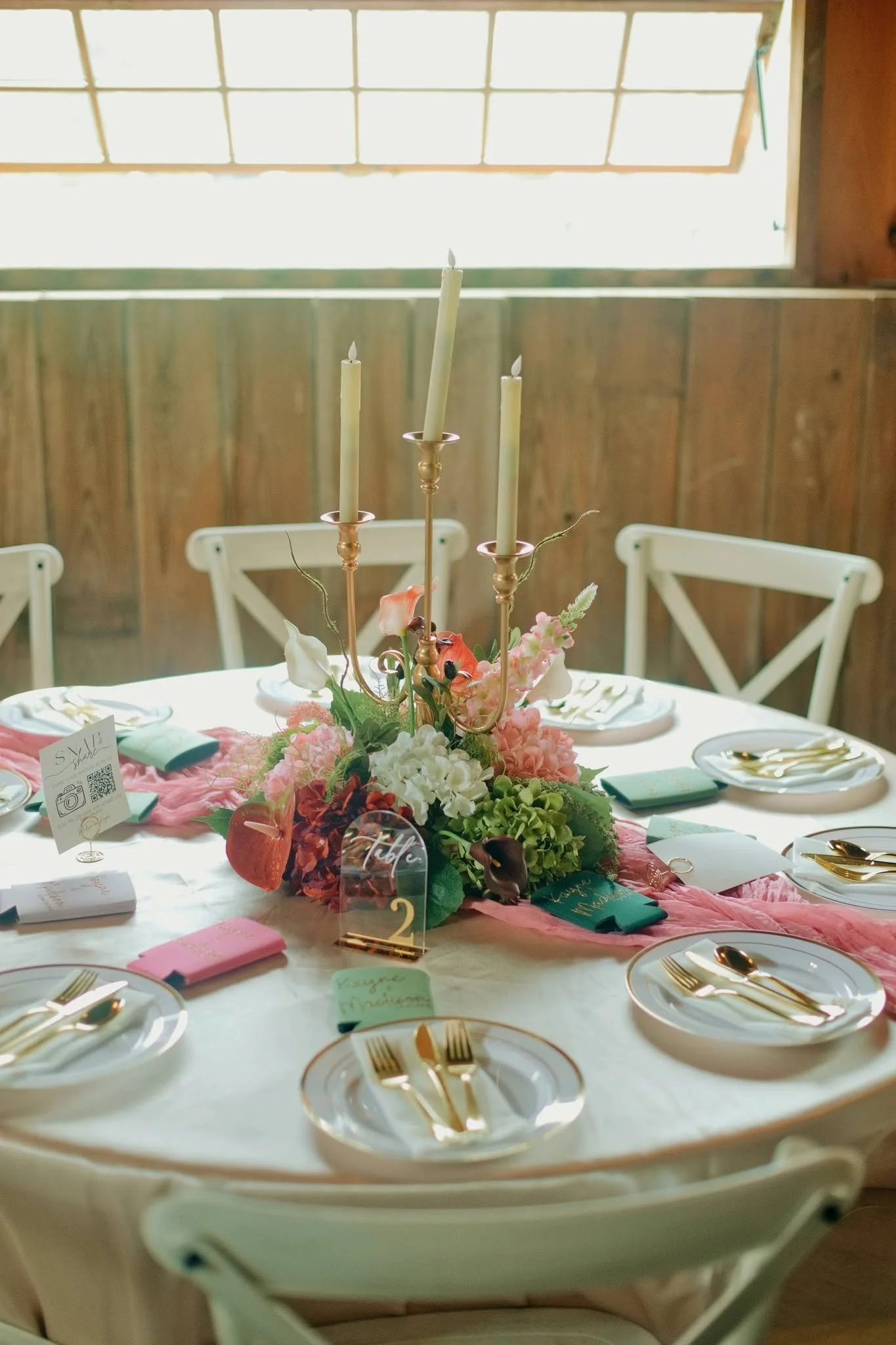 Decorated wedding reception table with floral centerpiece, gold candlesticks, plates, gold cutlery, pink napkins, and place cards, set for a special occasion or celebration.