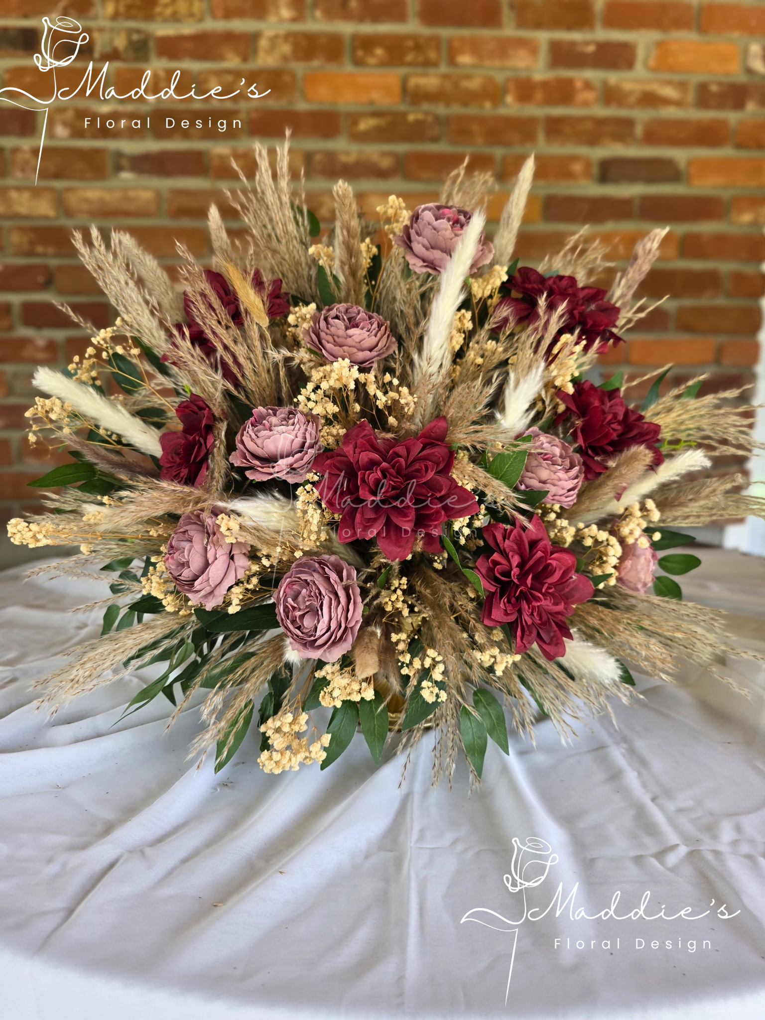 A floral arrangement with pink and maroon flowers, dried pampas grass, and green leaves, displayed on a white fabric surface with a brick wall background.