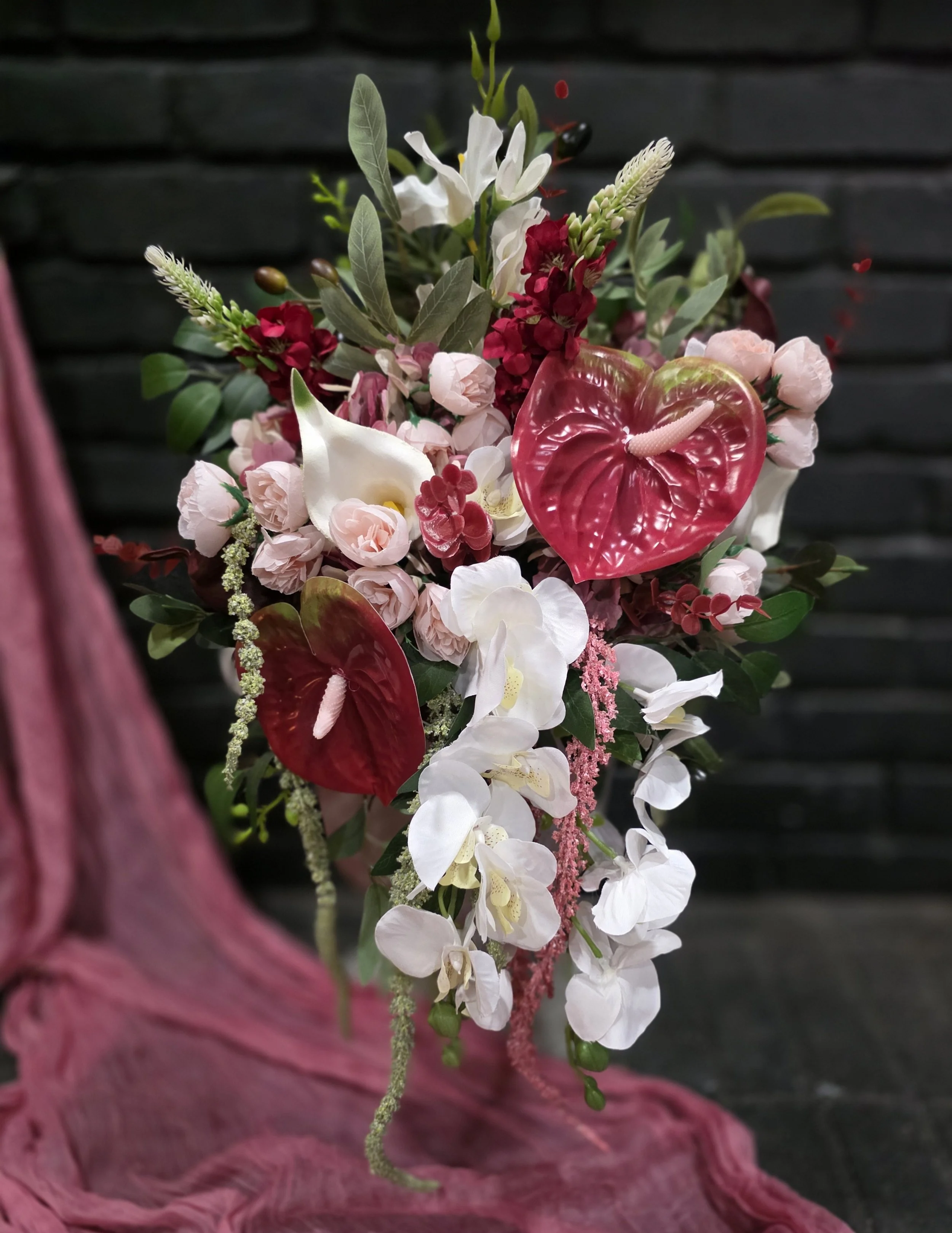A colorful flower arrangement with white, pink, and red flowers, green leaves, and decorative elements, placed on a pink cloth against a dark background.