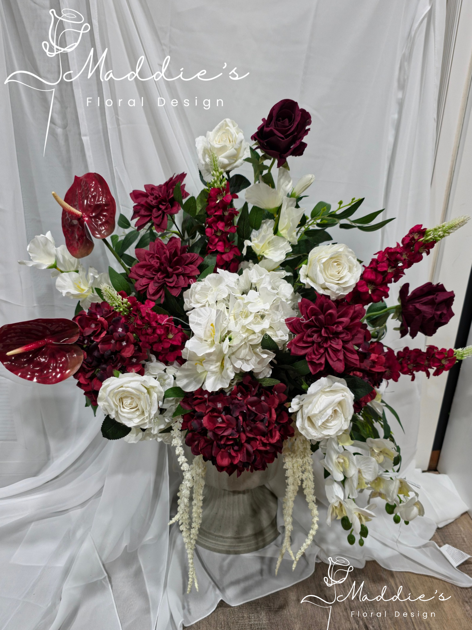 A floral arrangement with white roses, burgundy roses, and red and white anthuriums, set against a white sheer backdrop.