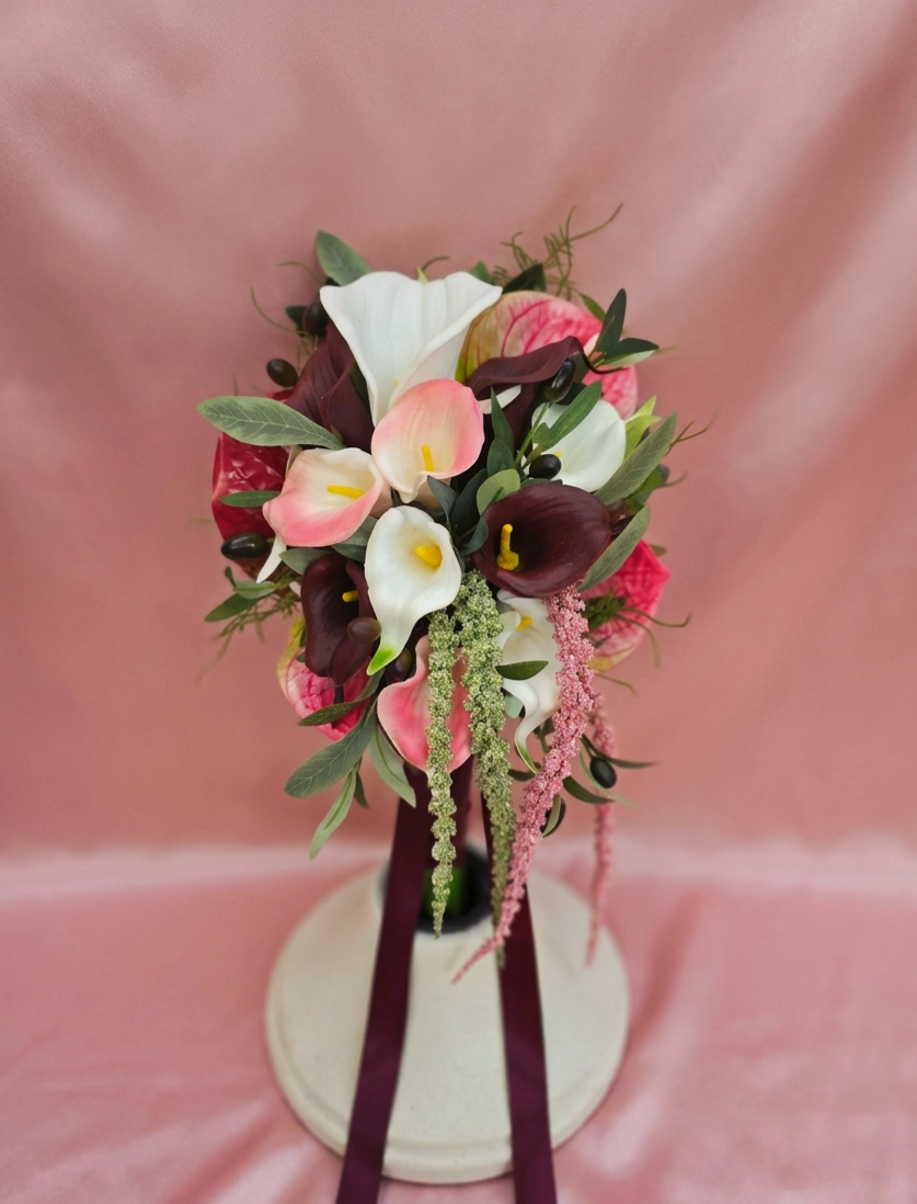 A floral bouquet with white, pink, and dark purple calla lilies, greenery, and hanging pink and green amaranthus, arranged in a vase with dark purple ribbons, set against a pink background.