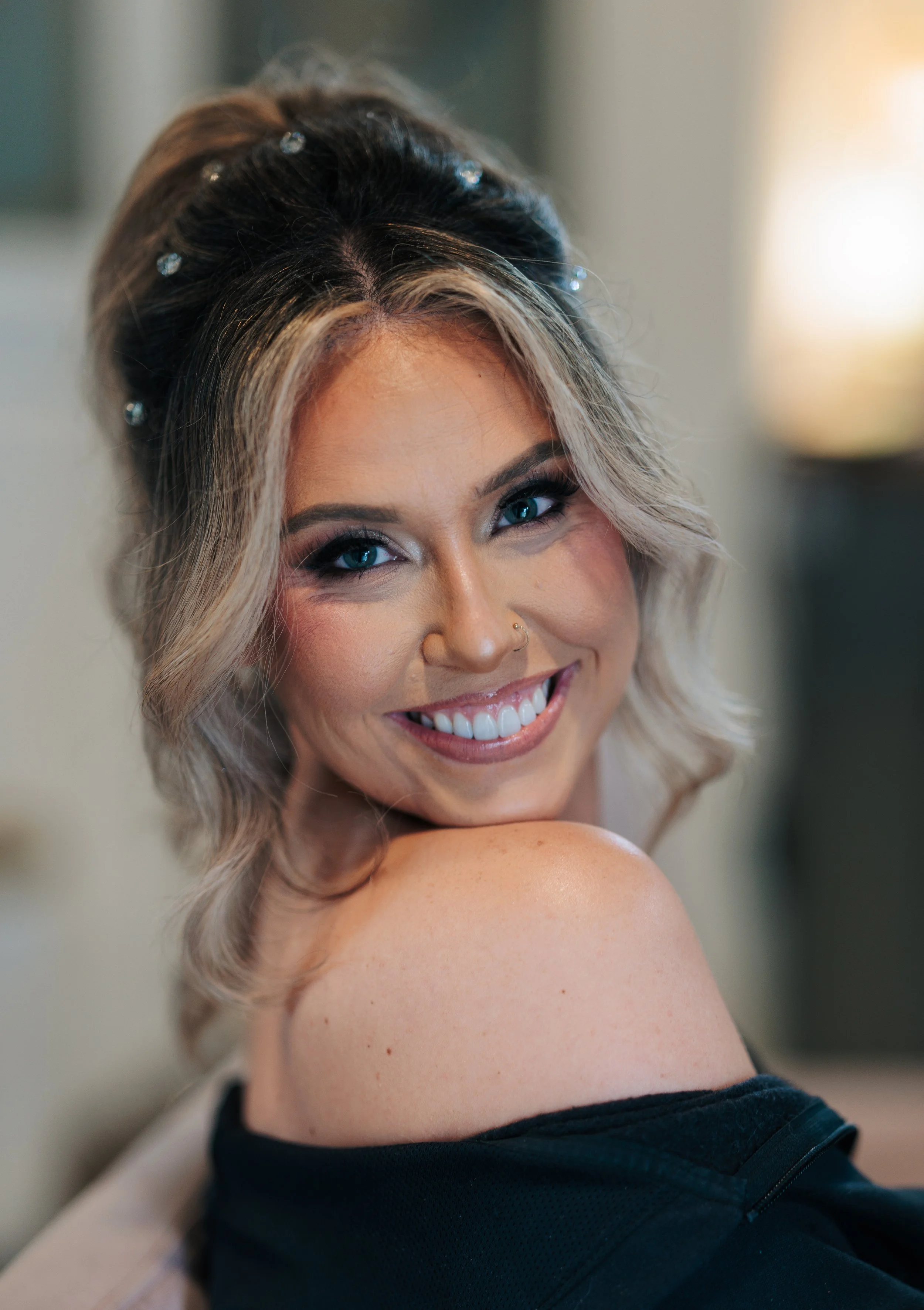 A woman with blonde hair styled in loose waves, shaved on one side, smiling and showing white teeth, wearing a black sleeveless top, and looking over her shoulder.