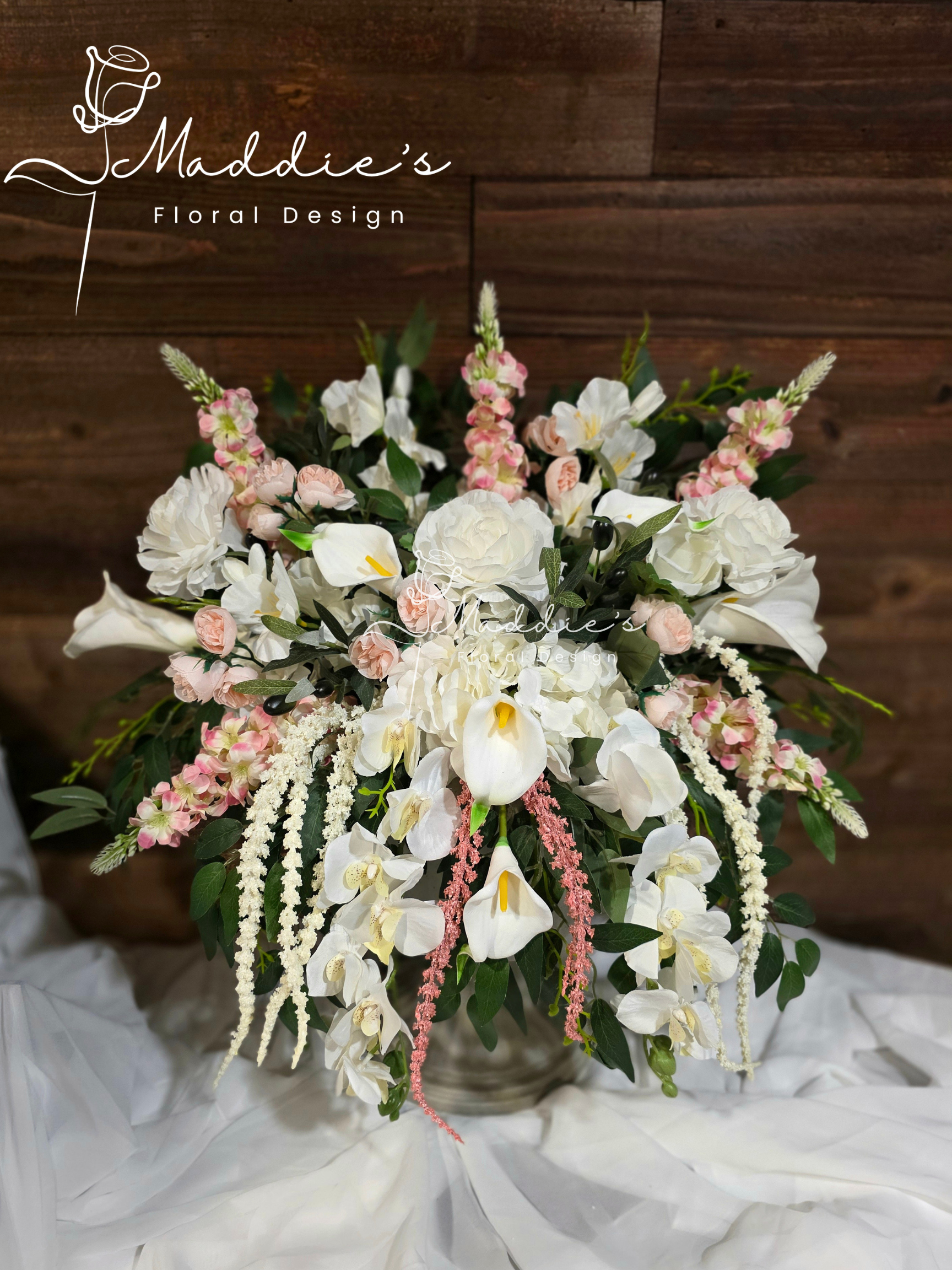 A wedding flower arrangement with white roses, calla lilies, pink flowers, and greenery in a white paper wrapping in front of a wooden wall.