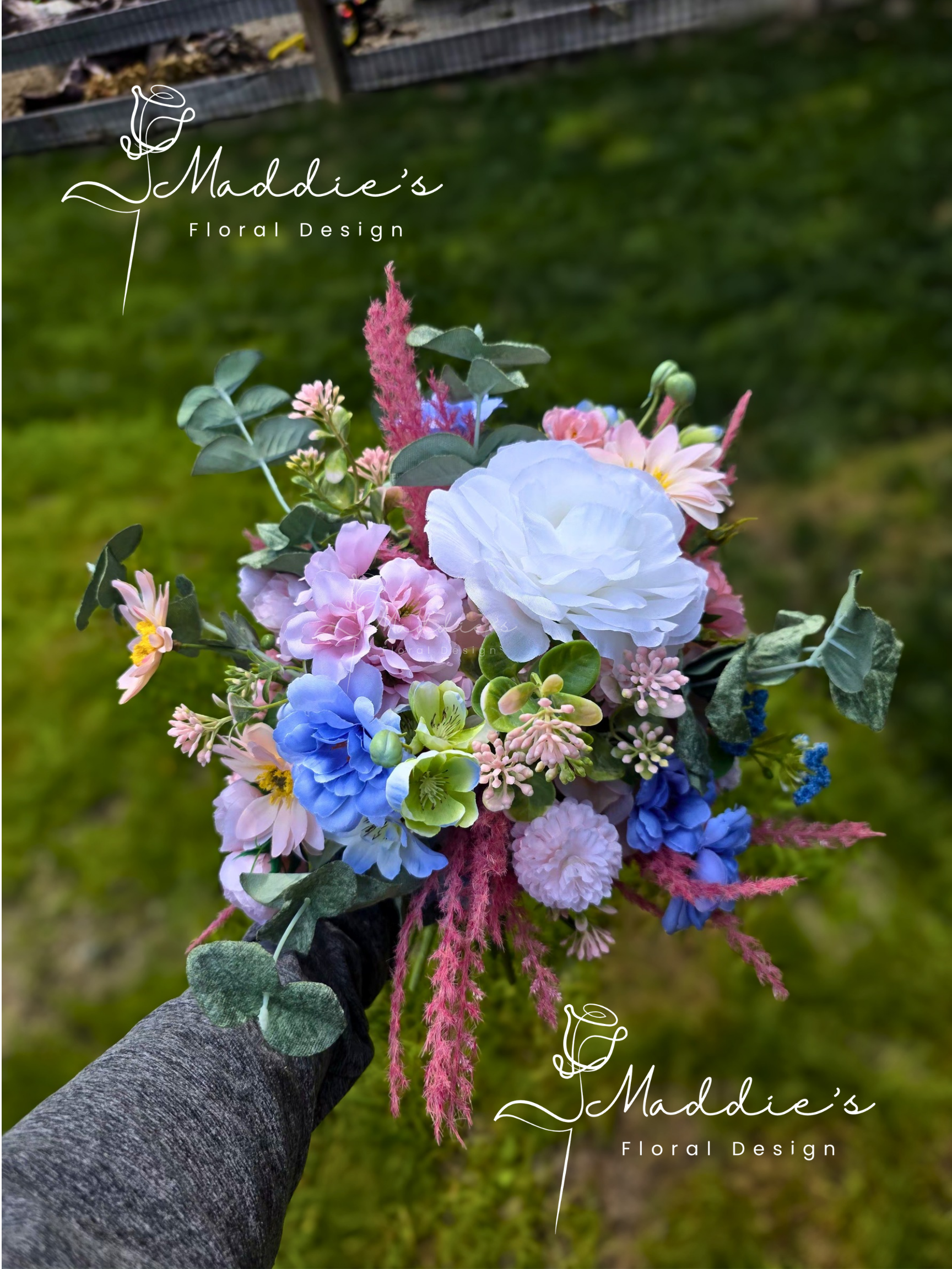 A florist's hand holding a vibrant bouquet of pink, purple, blue, and white flowers with greenery, outdoors on grass.