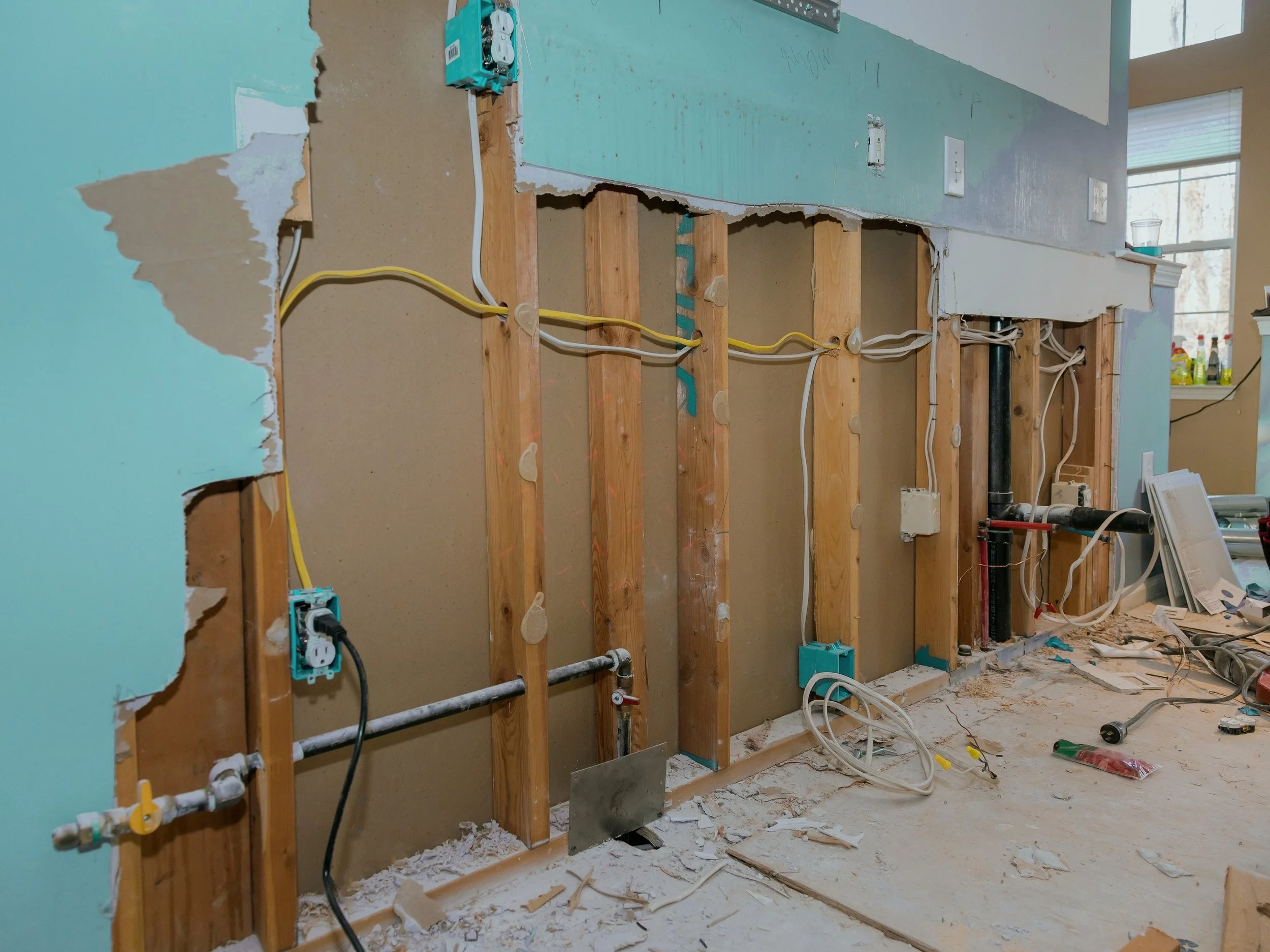A partially demolished interior wall with exposed wooden studs, electrical wiring, and plumbing pipes, indicating ongoing renovation work in a house.