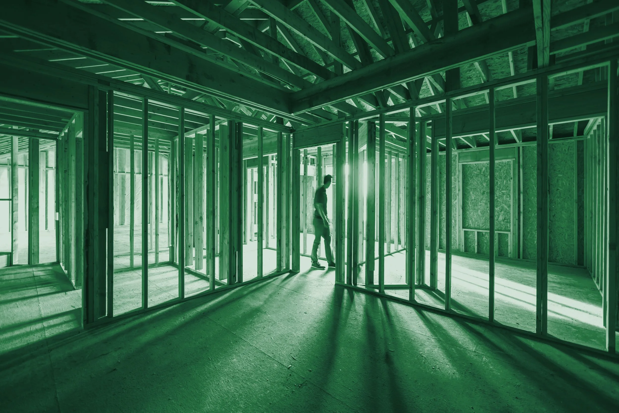 Construction site with wooden framing and a person walking inside the partially built structure, bathed in green light.