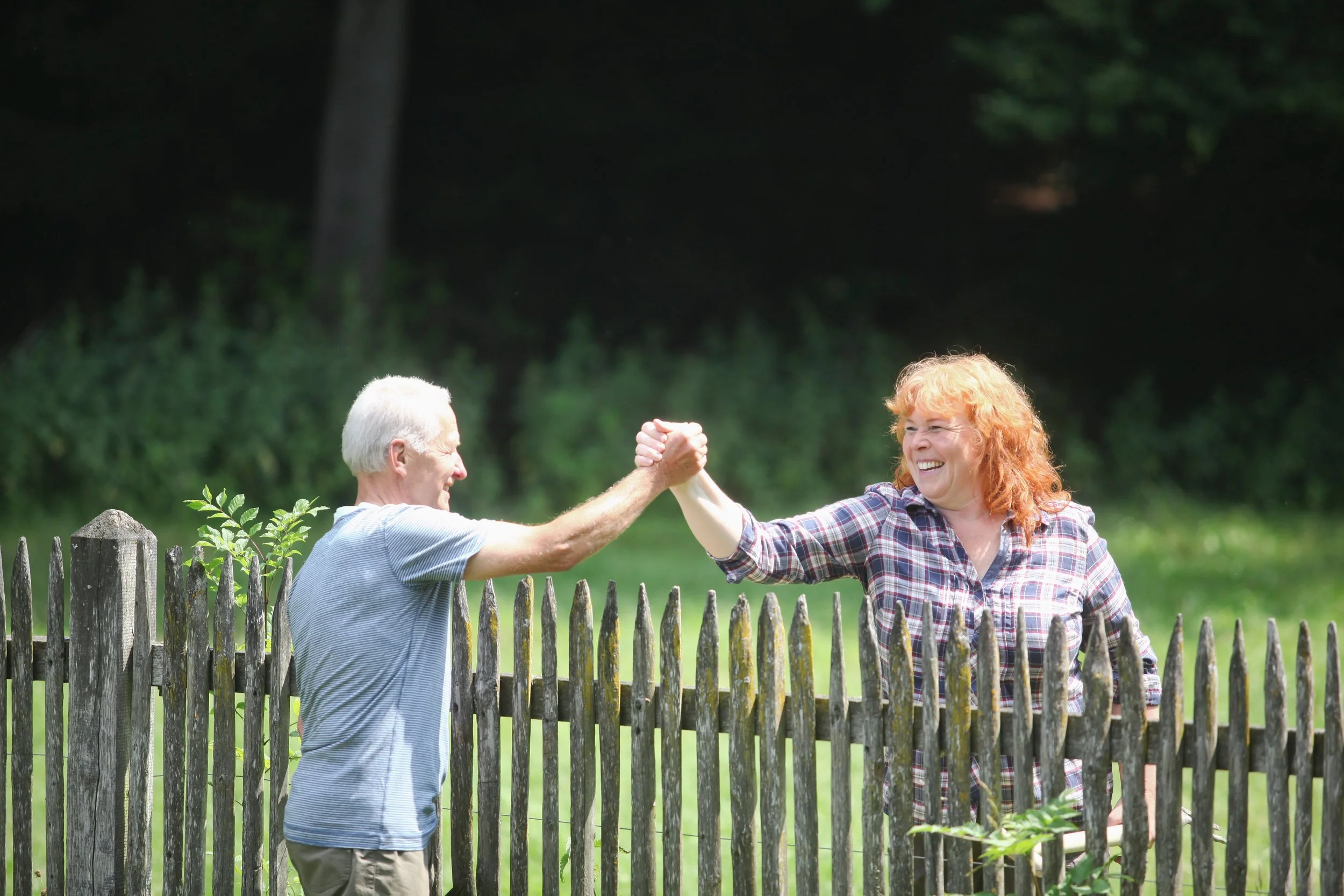 An elderly man and woman are smiling and engaging in a handshake over a wooden fence in a green park, with trees in the background.