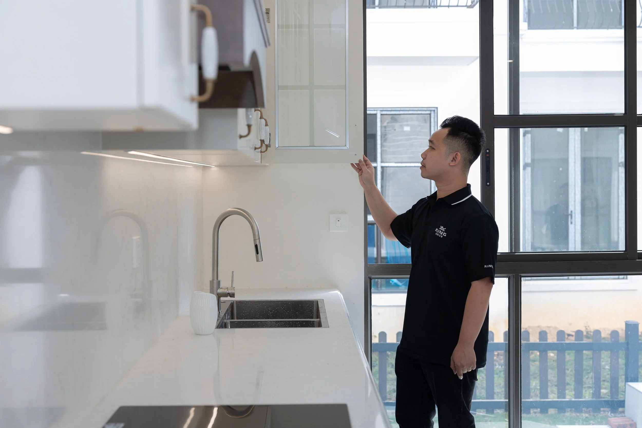 A man in a black uniform looks at a kitchen cabinet in a modern kitchen with large windows.