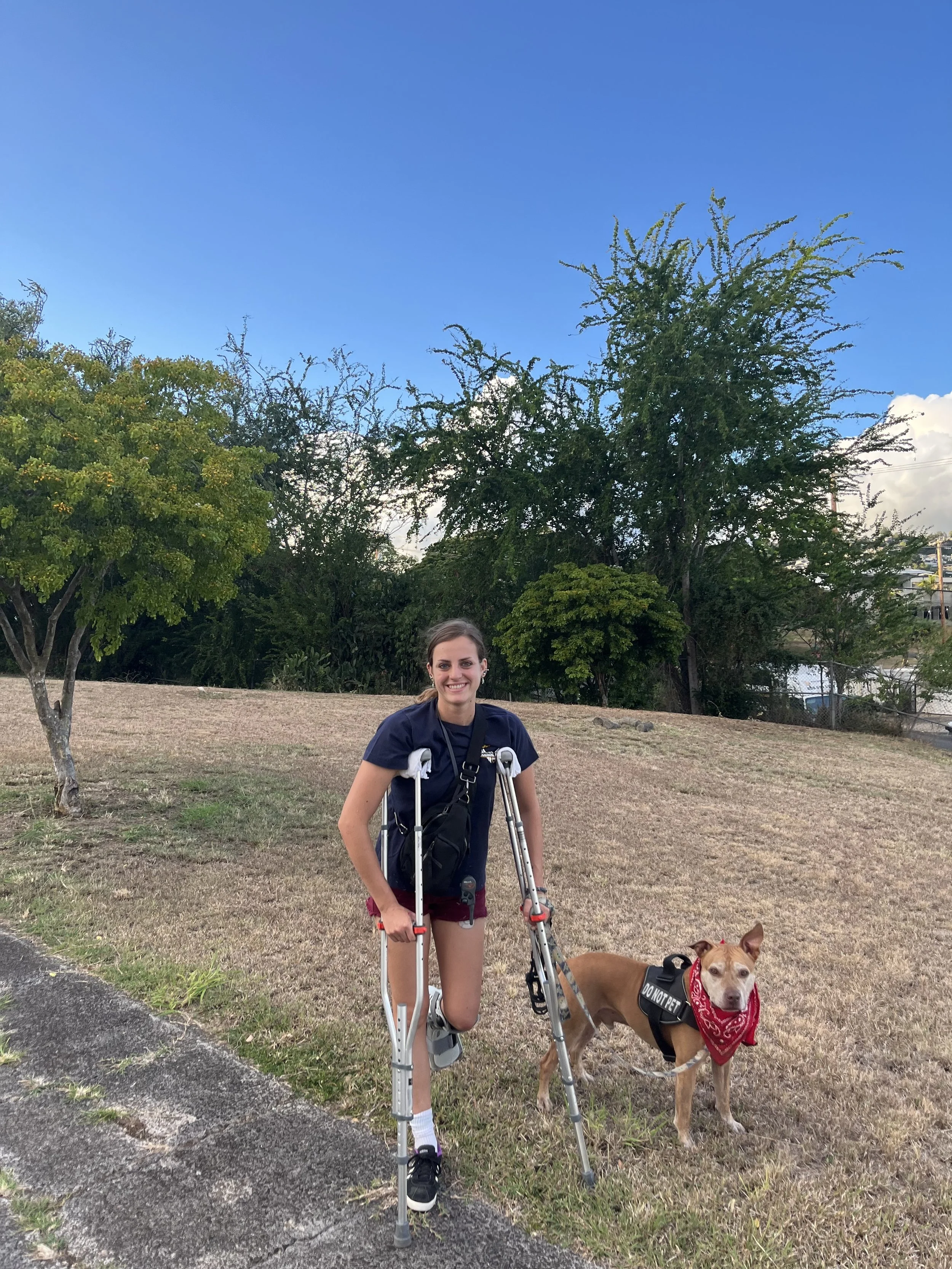 A young woman with a prosthetic leg standing outdoors on a grassy area with trees in the background, smiling at the camera, with a brown pitbull dog wearing a 'DO NOT PET' vest and a red bandana around its neck.