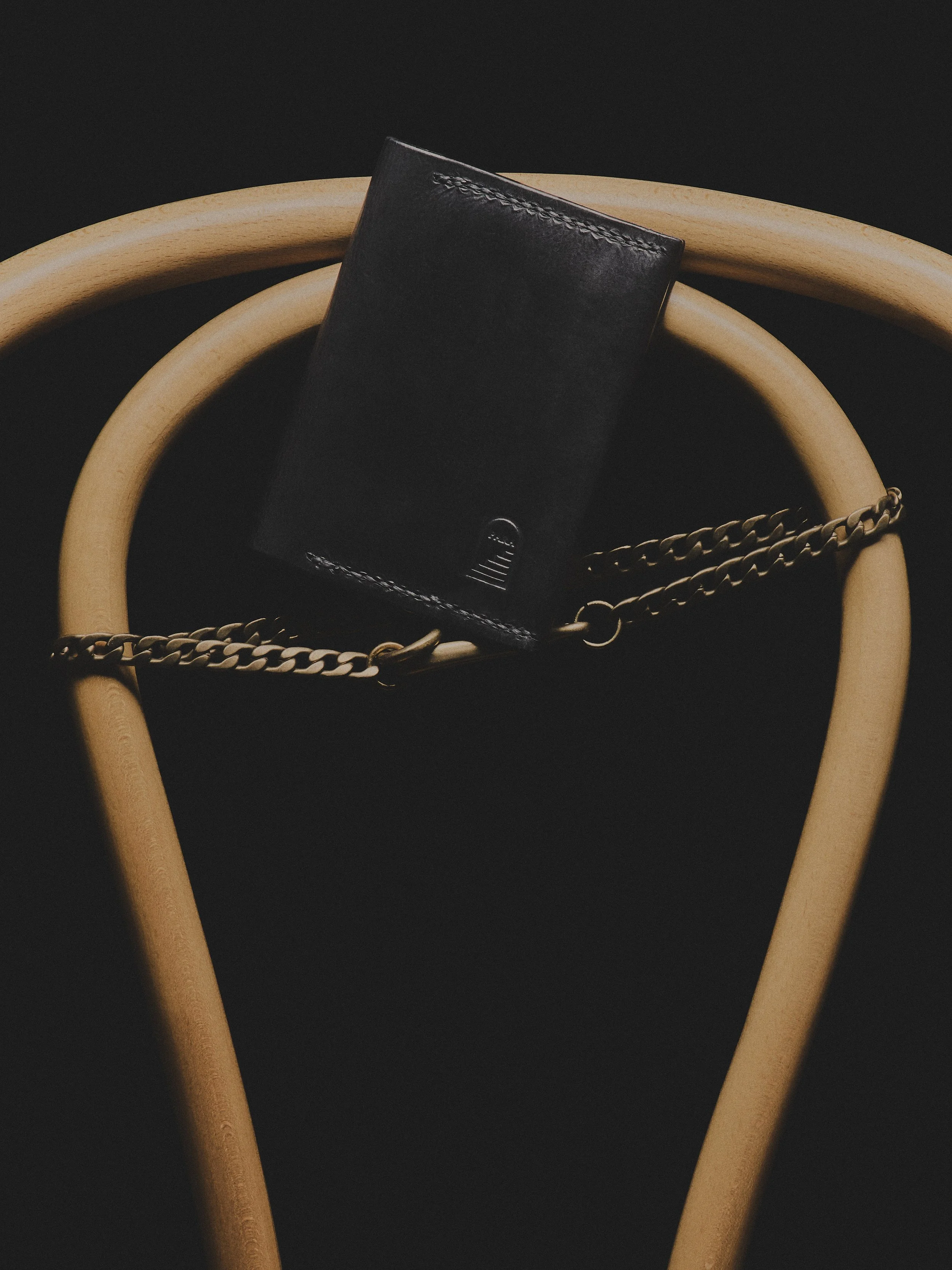 A black leather wallet with stitching on the top edge positioned between two circular wooden hoops, with a chain resting on one hoop, all against a dark background.