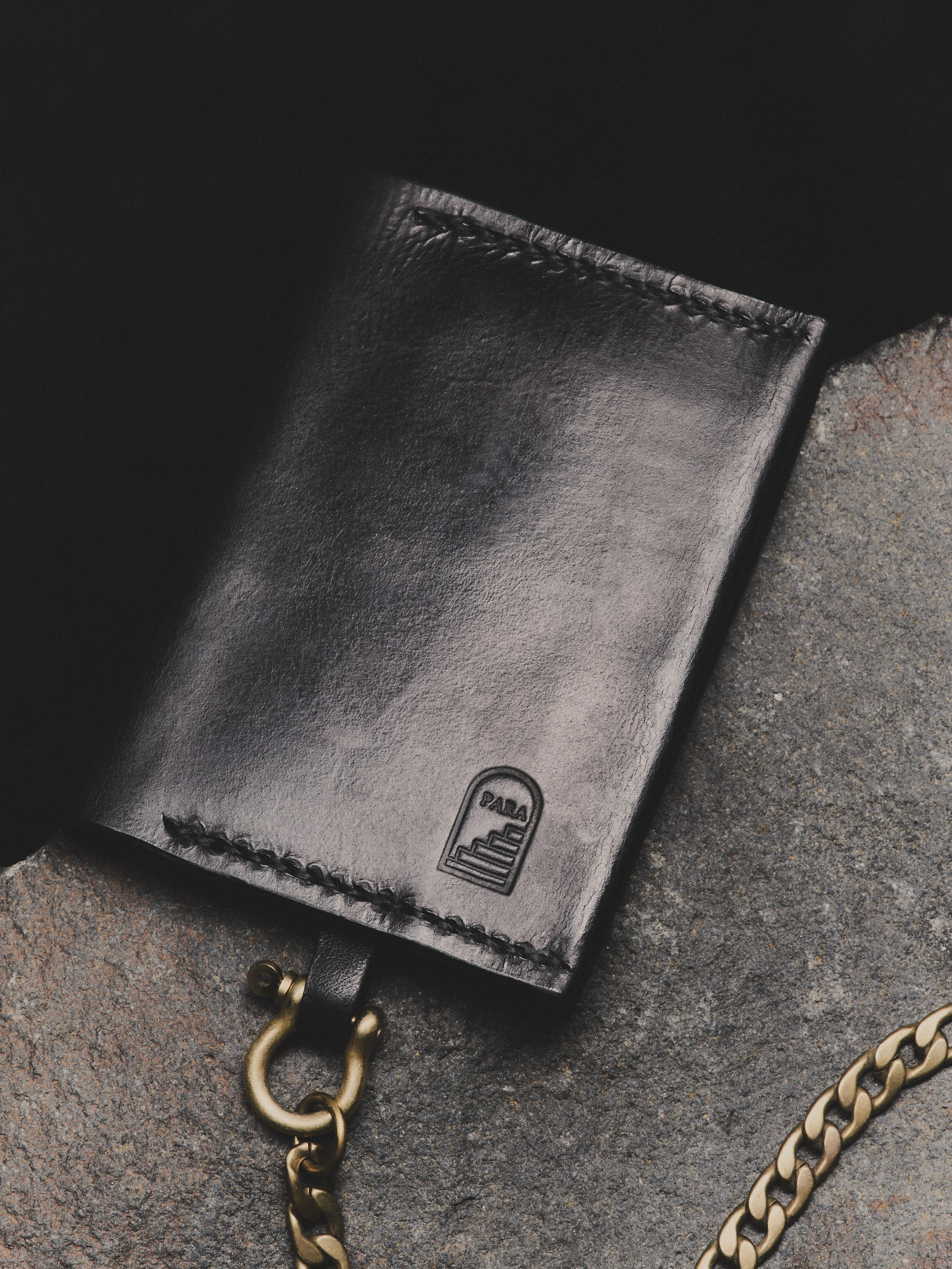 Close-up of a black leather wallet with a small logo embossed in the corner, placed on a textured surface, with a brass chain attached to it.