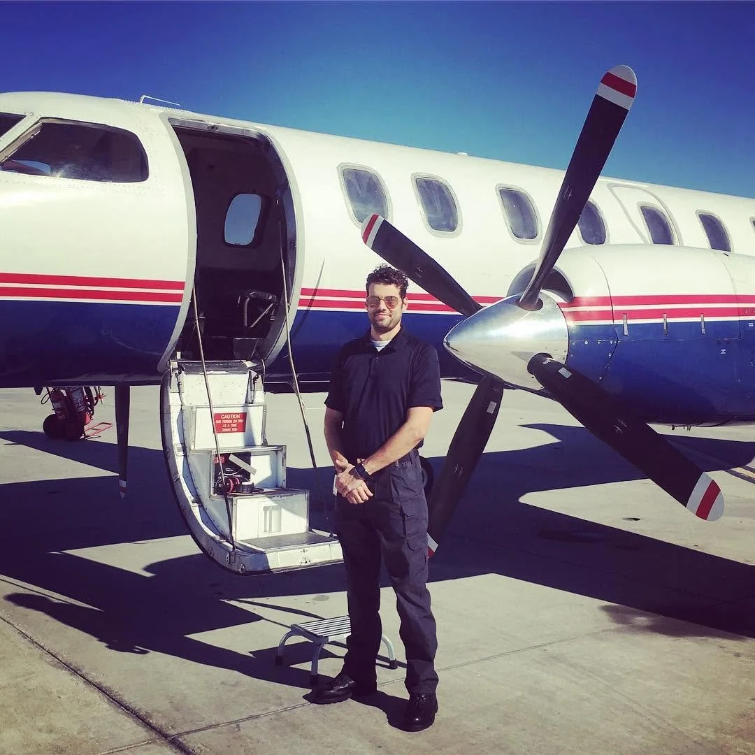 Man standing at the entrance of a twin-engine, turbo-propeller airplane, on a tarmac, against a clear blue sky. This was the first airline position worked by Max Schwartz, as a first officer, at a company called "Berry Aviation." Aircraft was SA-227.