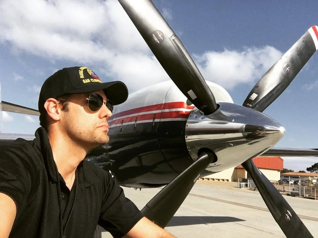 Man wearing Rayban Aviator sunglasses and a US NAVY cap (with a military patch.) He took this selfie sitting on the steps of the aircraft, under a partly cloudy sky. The aircraft in reference is the "FairChild Metro-Liner" (SA-227)