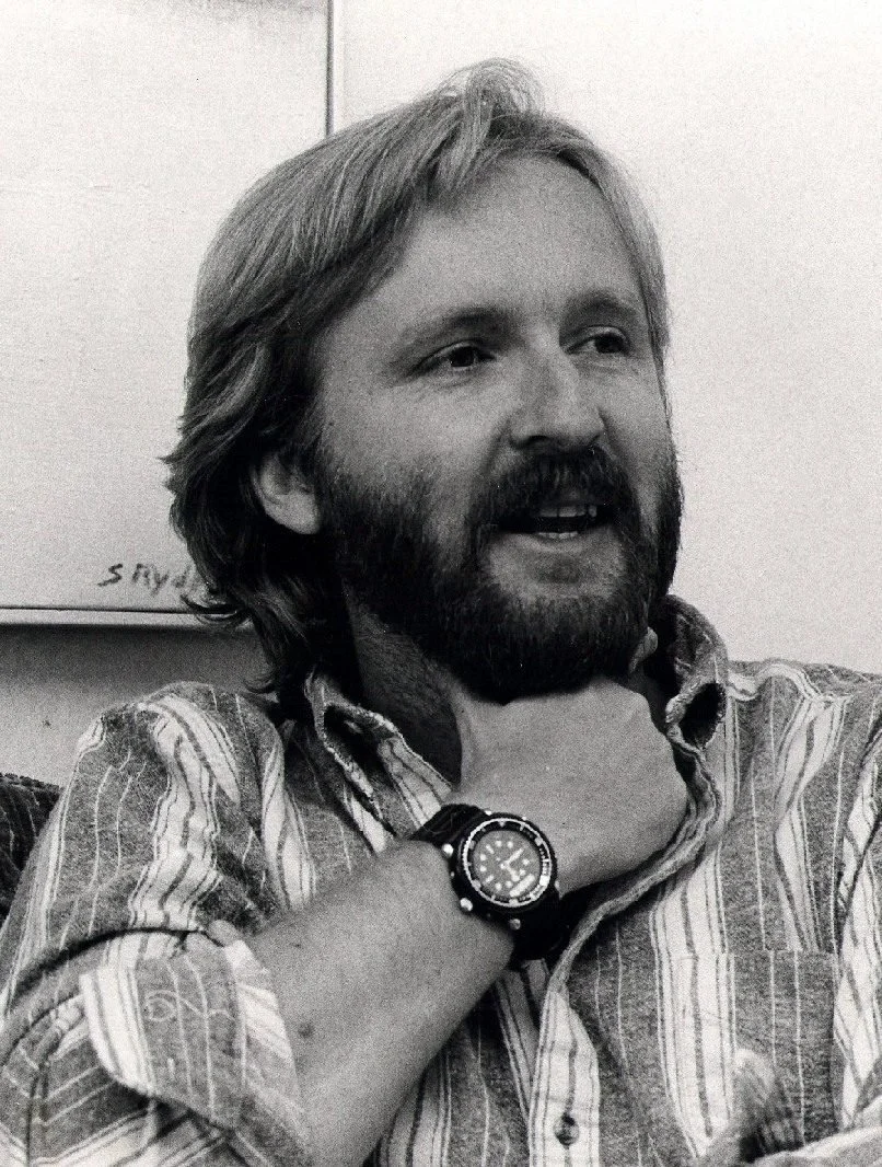 Young James Cameron, with long hair and a beard, wearing a striped shirt and a wristwatch, sitting indoors and smiling. He knows  deep down he's going to create many of the 20th century's most iconic films.