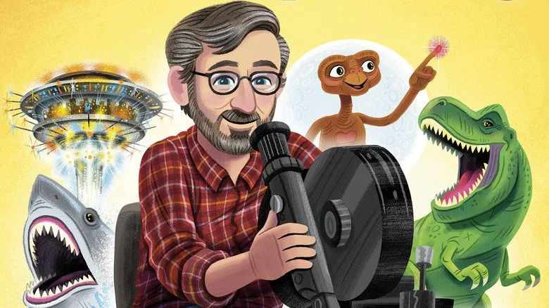 Illustration of Steven Speilberg, with glasses and beard, operating a camera, surrounded by a flying spaceship, a friendly T-Rex, a shark with open mouth, and smiling E.T. -- with a raised finger emitting his heart light, against a yellow background.
