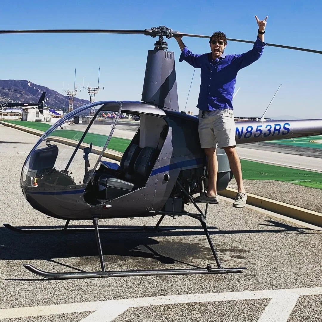 A man -- Max Schwartz -- with dark curly hair, sunglasses, a blue button-up shirt, and shorts. He's doing a pre-flight inspection on a Robinson 22 (R-22) helicopter, smiling and making a peace sign at the camera. The R-22 is an economical trainer.