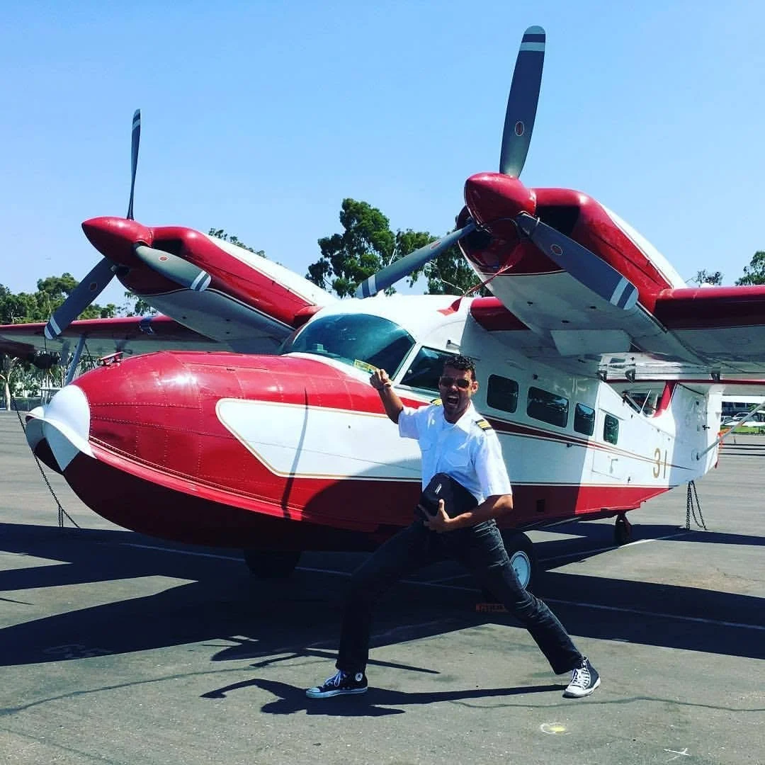 Max Schwartz in his pilot's uniform, posing excitedly in front of a red and white amphibious airplane on the tarmac. This was a "Grumman Widgeon," the same make and model which he later earned his Multi-Engine Seaplane rating in.