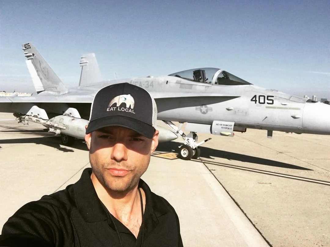 Max Schwartz wearing a cap that says "Eat Local." Taking a self-portrait in front of an F-18 Super Hornet fighter jet. Some trivia for you. Max's boss forced him to stop wearing the hat, because it conflicted with Navy / company policies.