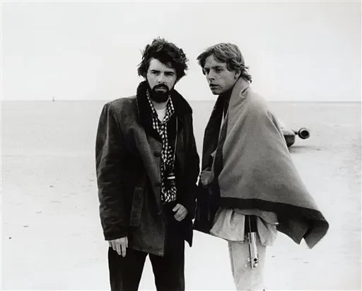 Two men standing on a beach in black and white, wearing coats and scarves, looking serious at the camera.