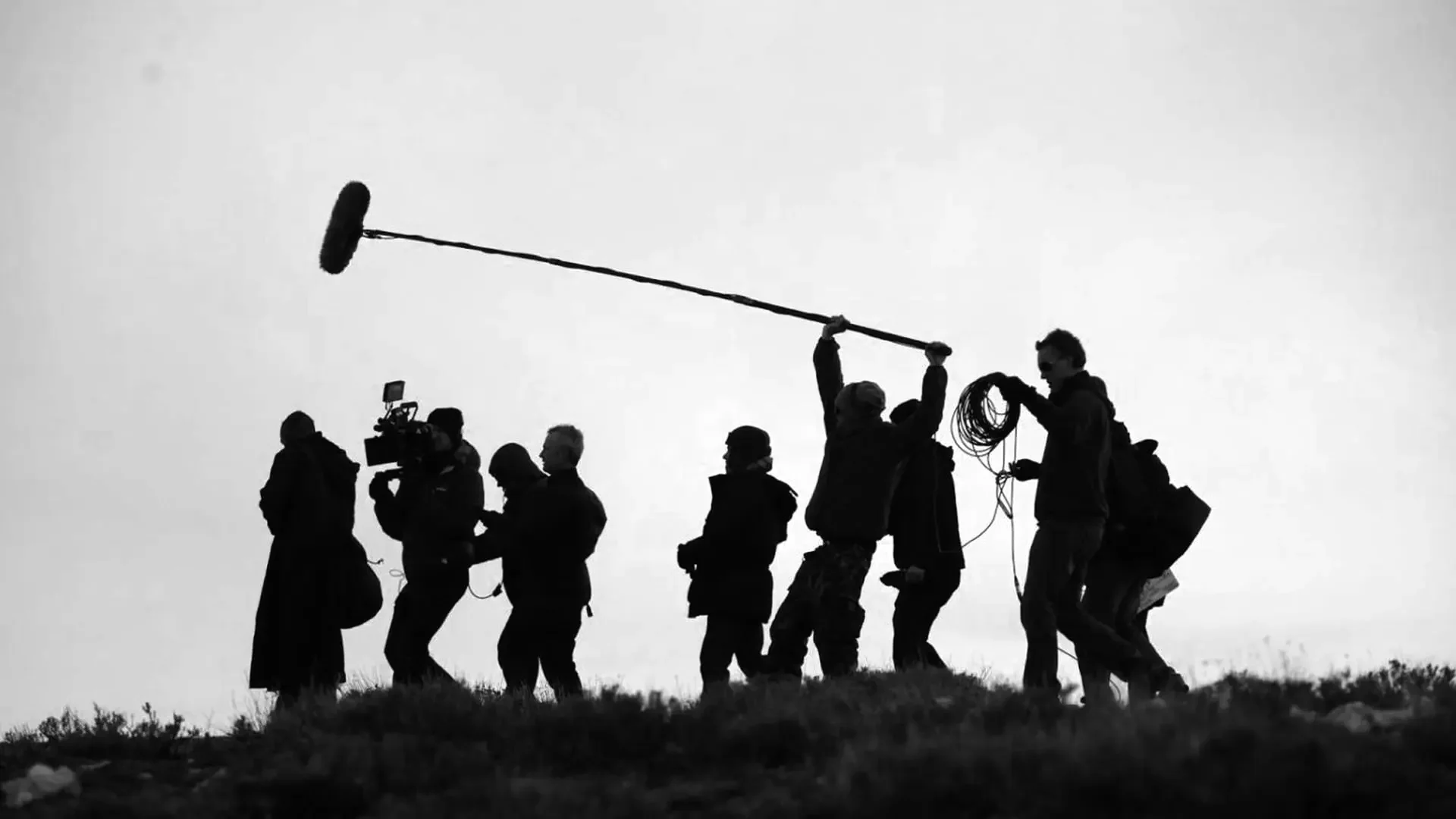 Black and white silhouette of a small film crew, on a hill, making a movie. They are holding various pieces of audio and visual equipment, with one person extending a classic boom microphone overhead.