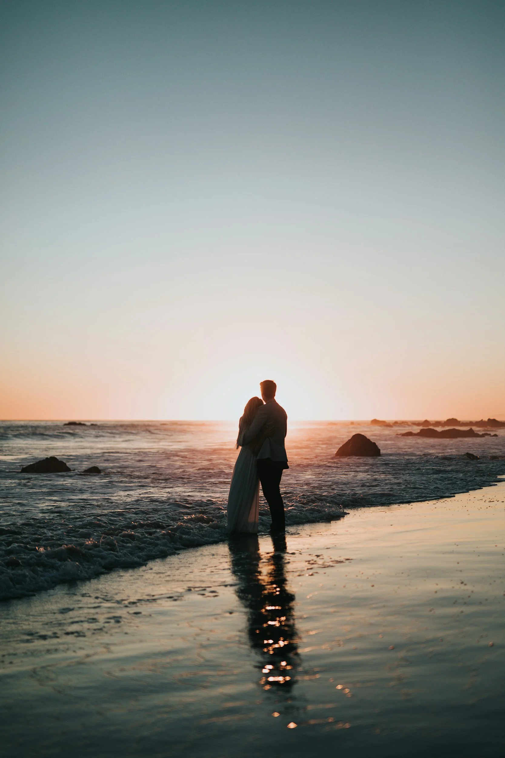 A couple stands on the beach at sunset, embracing and looking at the ocean.