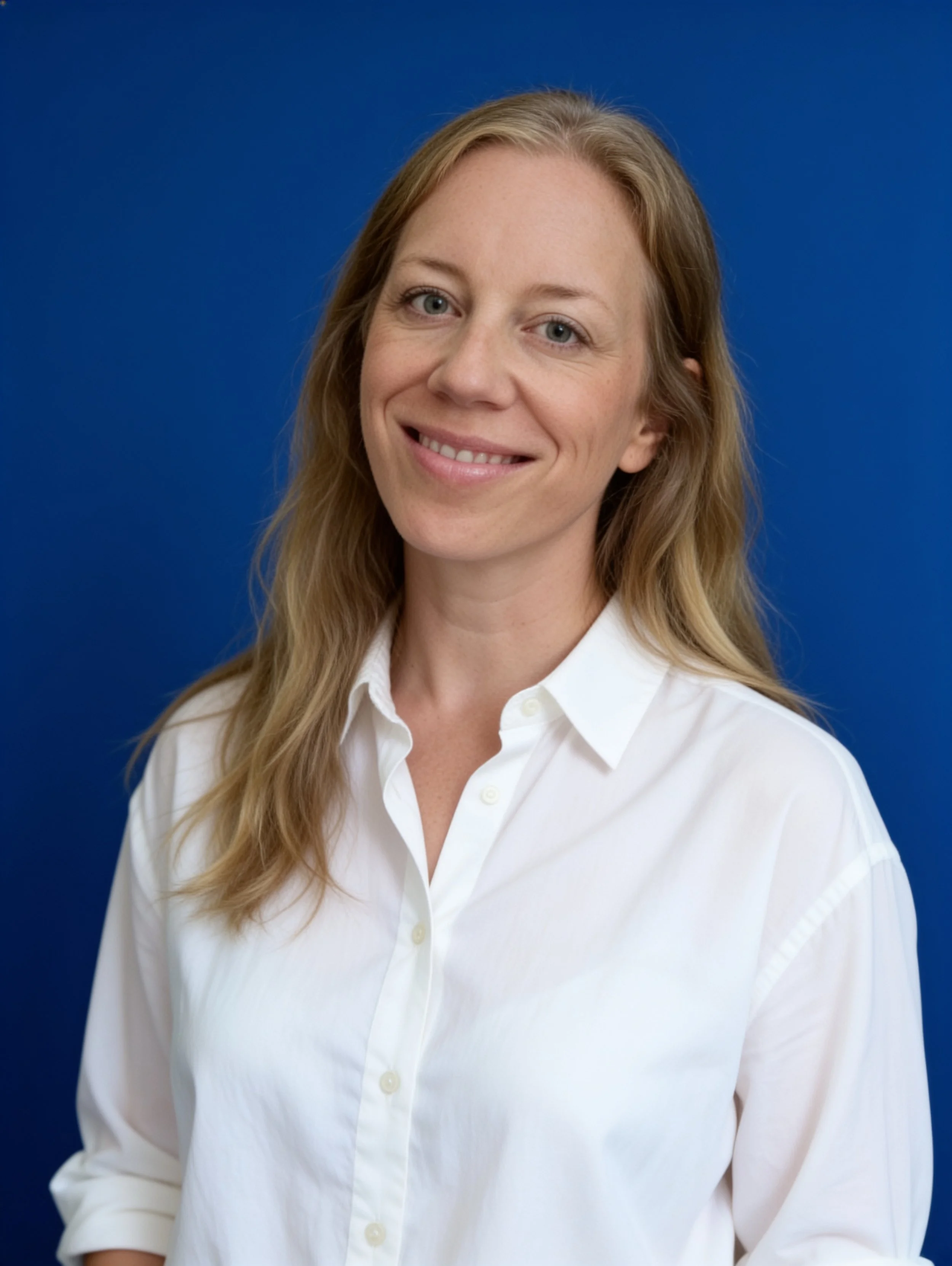 Portrait of a woman with long blonde hair, wearing a white button-up shirt, smiling in front of a solid blue background.