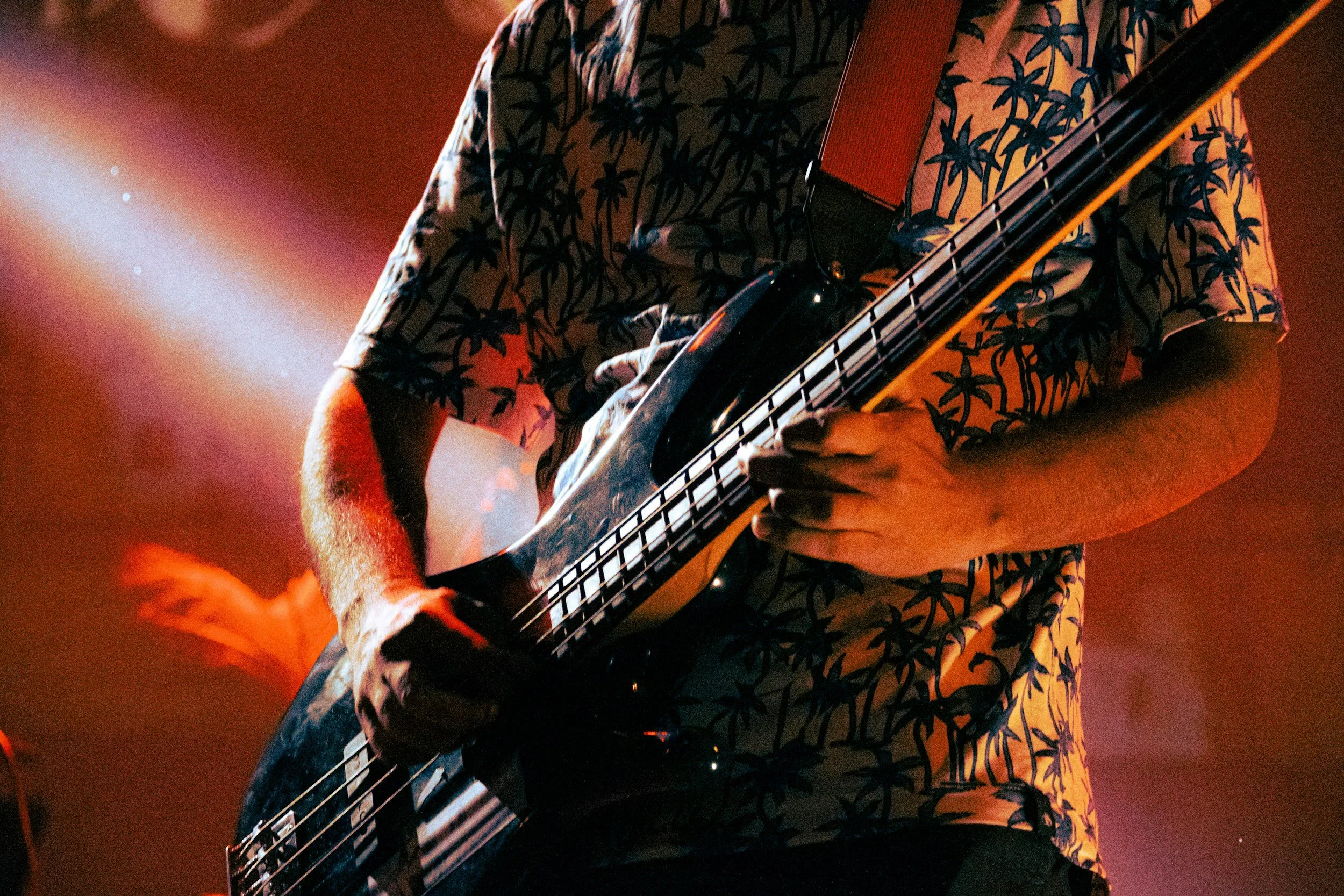 Person wearing a shirt with a palm tree pattern playing an electric bass guitar on stage with a red and orange background.