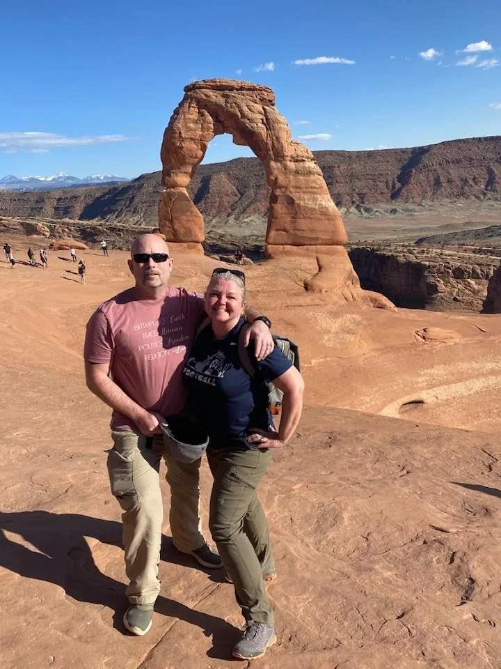Two people standing in front of the Delicate Arch, a natural sandstone arch, in Arches National Park, Utah.