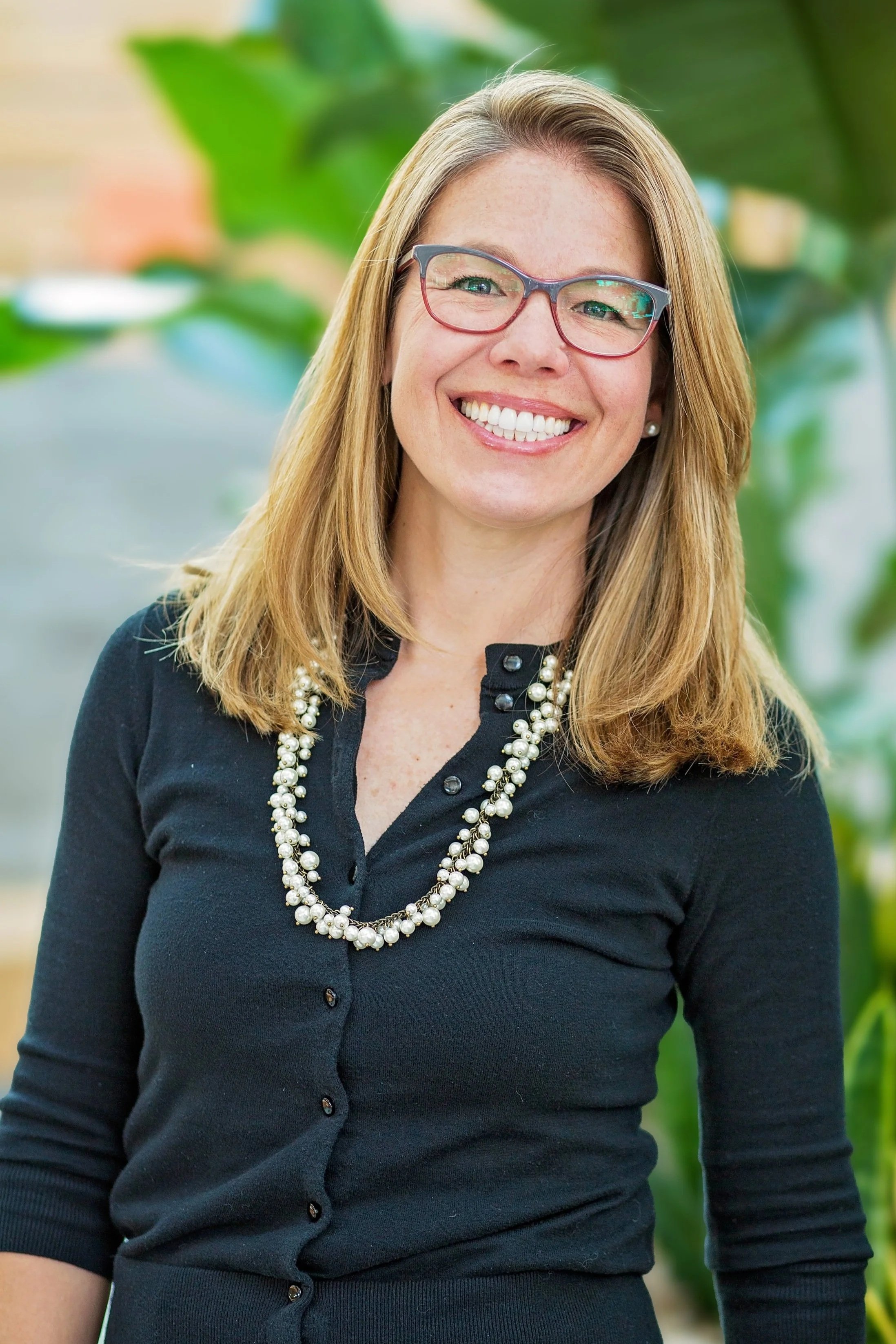A smiling woman wearing glasses, a black cardigan, and a pearl necklace standing outdoors with green plants in the background.