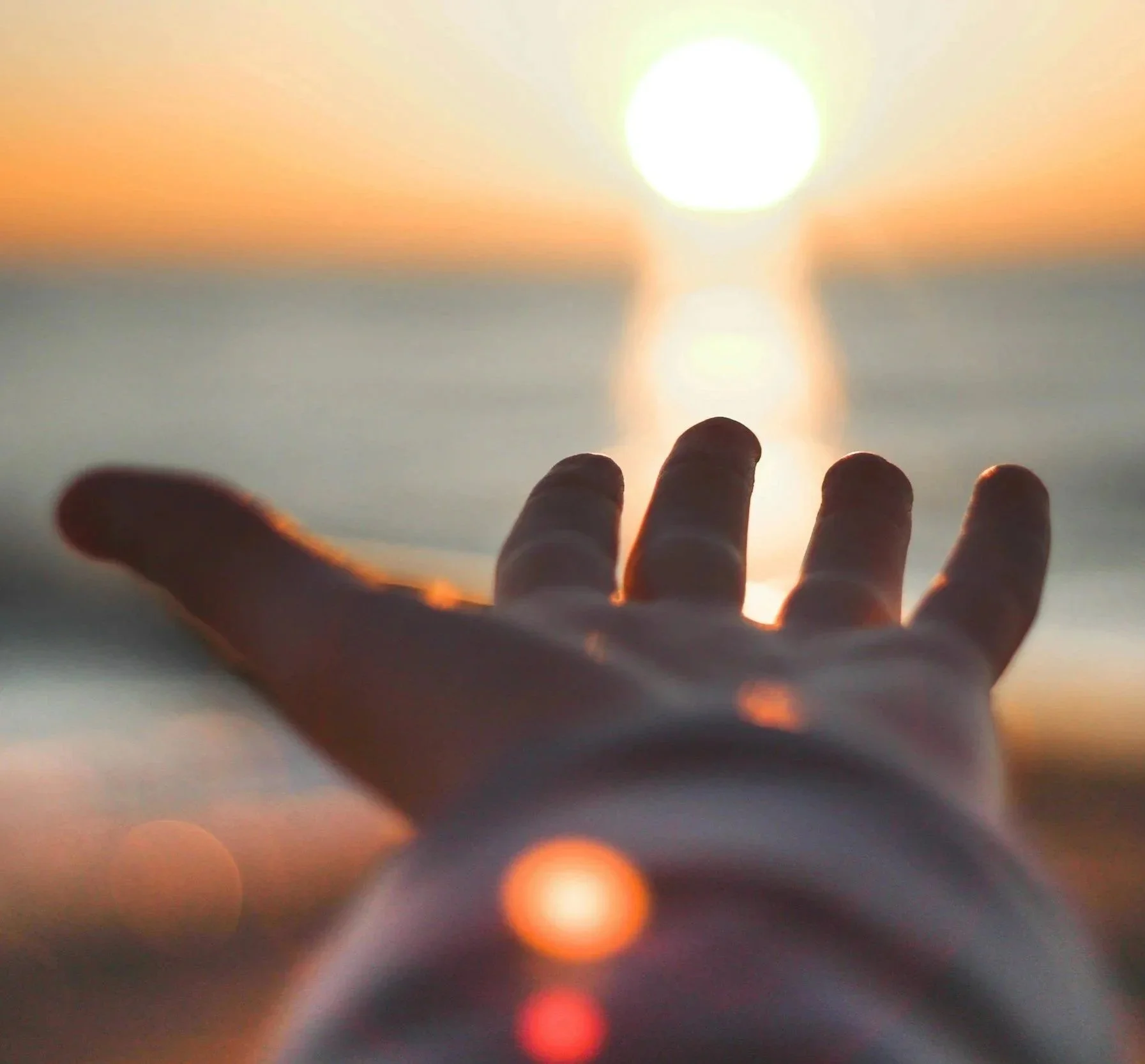 Close-up of a hand reaching out towards a bright sun over the ocean at sunset.