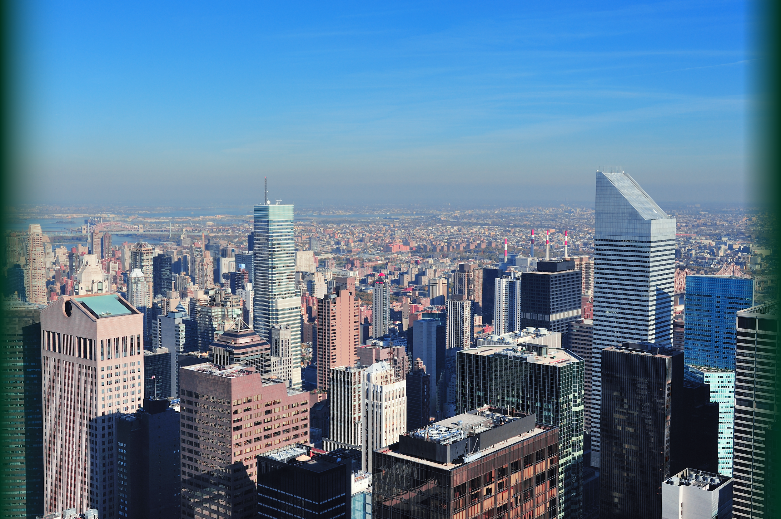 A city skyline with numerous high-rise buildings and skyscrapers under a clear blue sky. The photo appears to be taken from a tall vantage point, showcasing a dense urban area.
