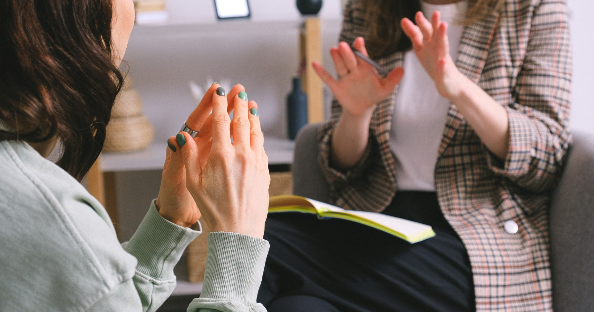 Two women having a conversation in a casual setting, one with hands clasped together and the other gesturing with a smartphone, with a notebook open.