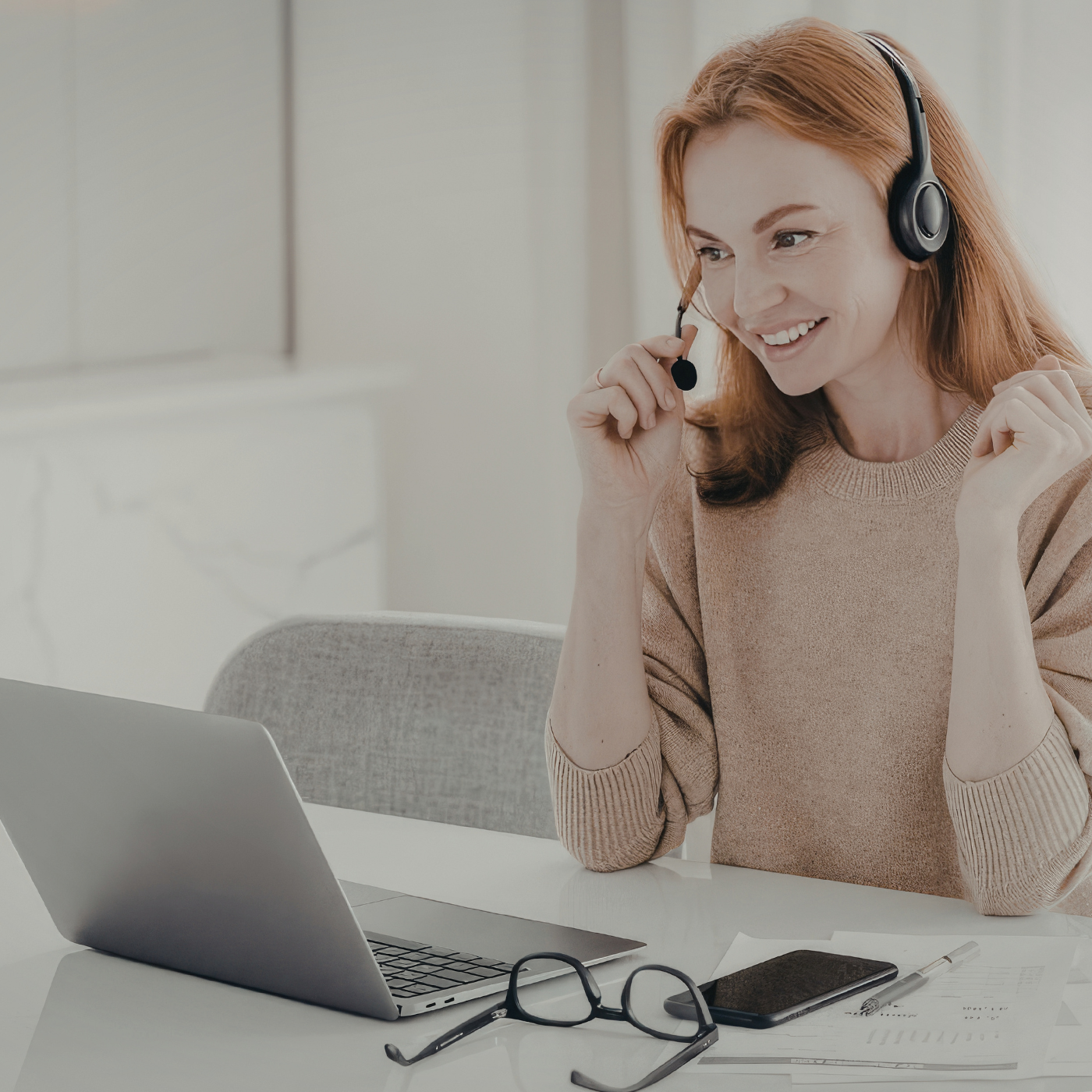 A woman with red hair wearing a beige sweater and a headset working at a desk with a laptop, glasses, smartphone, and papers.