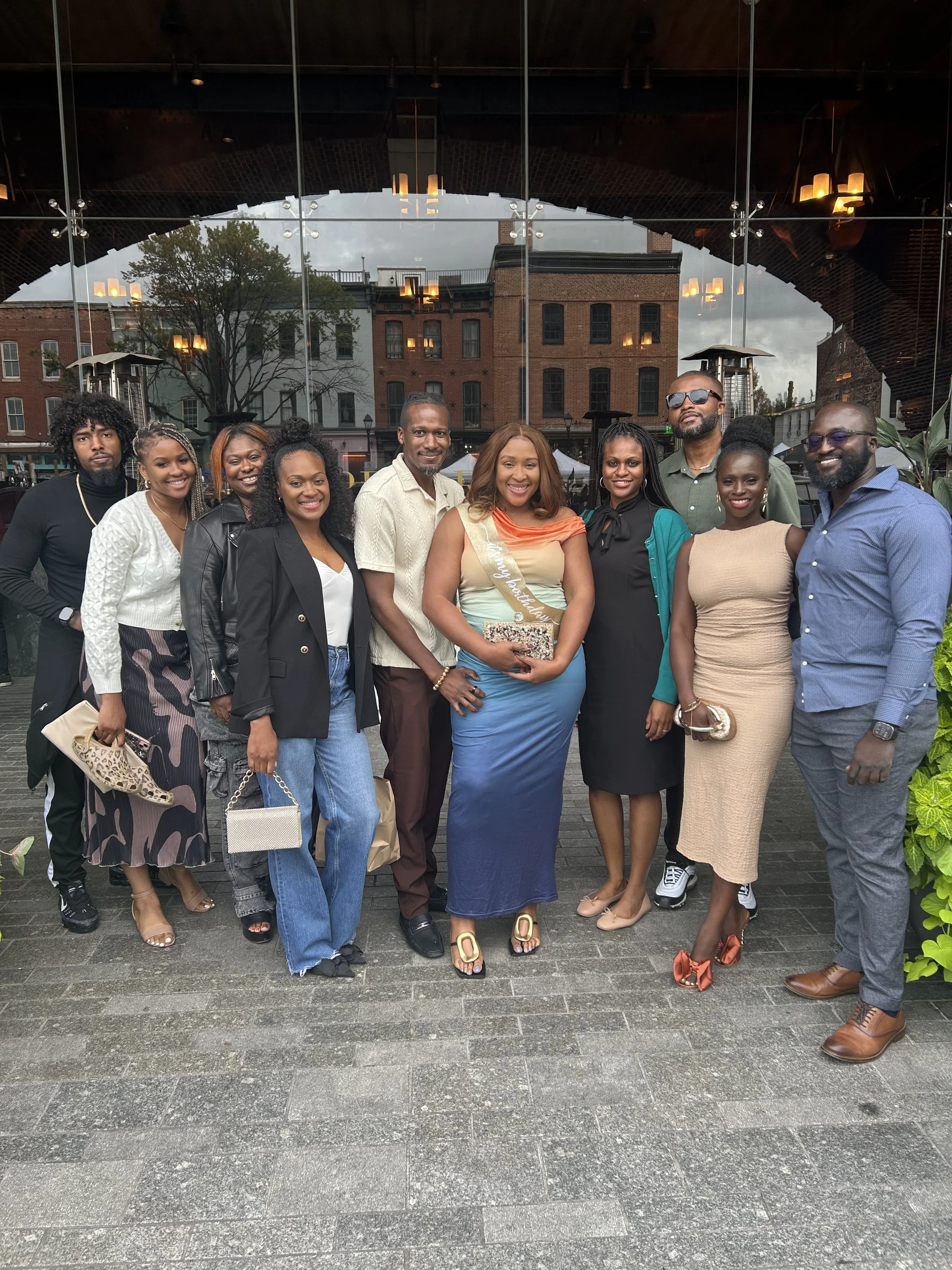 A diverse group of 11 people gathered together outside in front of reflective glass, brick buildings, and cloudy sky, smiling for a photo, with some wearing casual and semi-formal attire, and one woman wearing a sash that says 'Happy Birthday'.