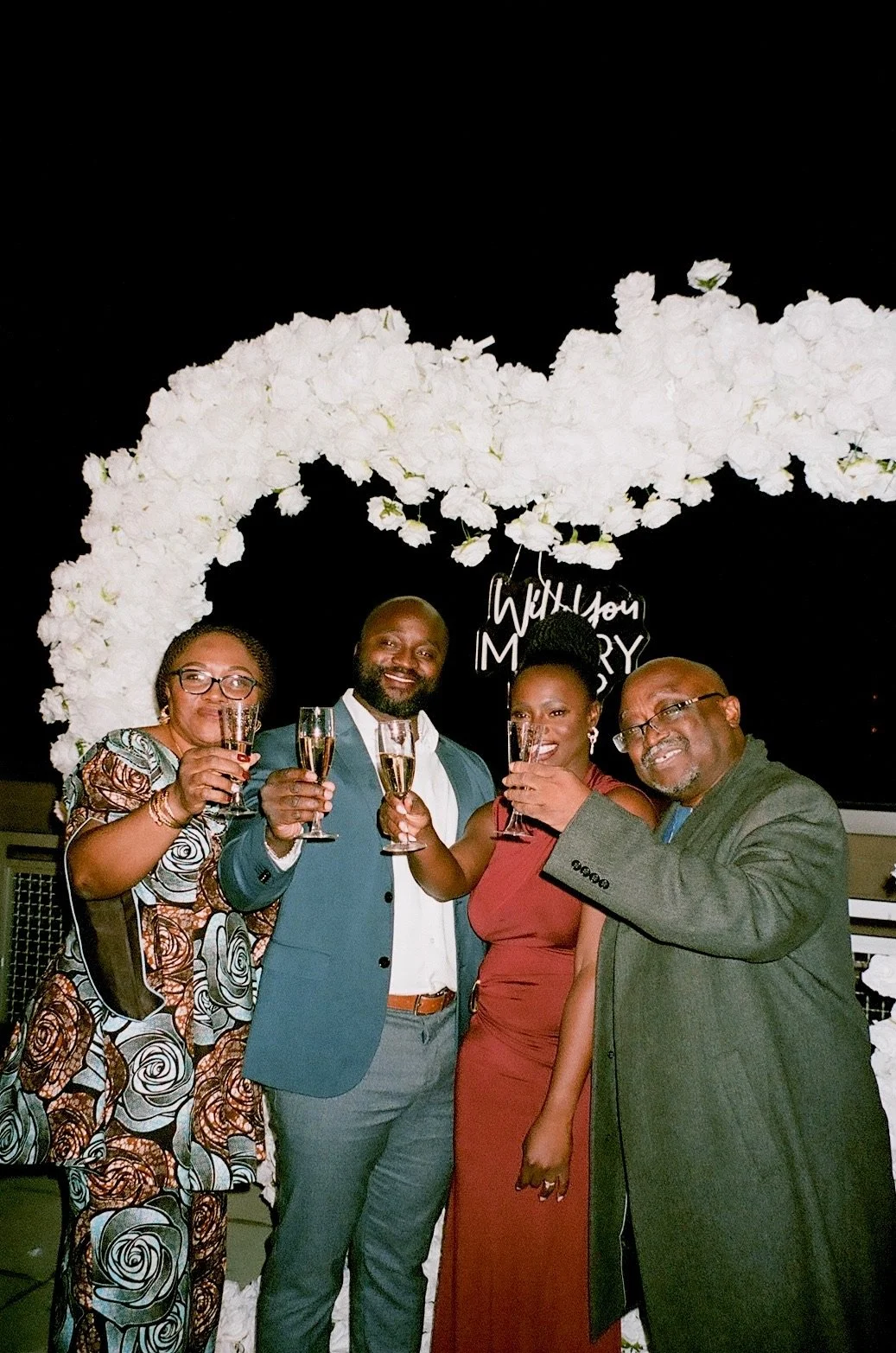 Moyo and Liam at their engagement celebration with their dads, standing under a white floral arch, holding glasses of champagne, with a sign that says 'Will You Marry?' in the background.