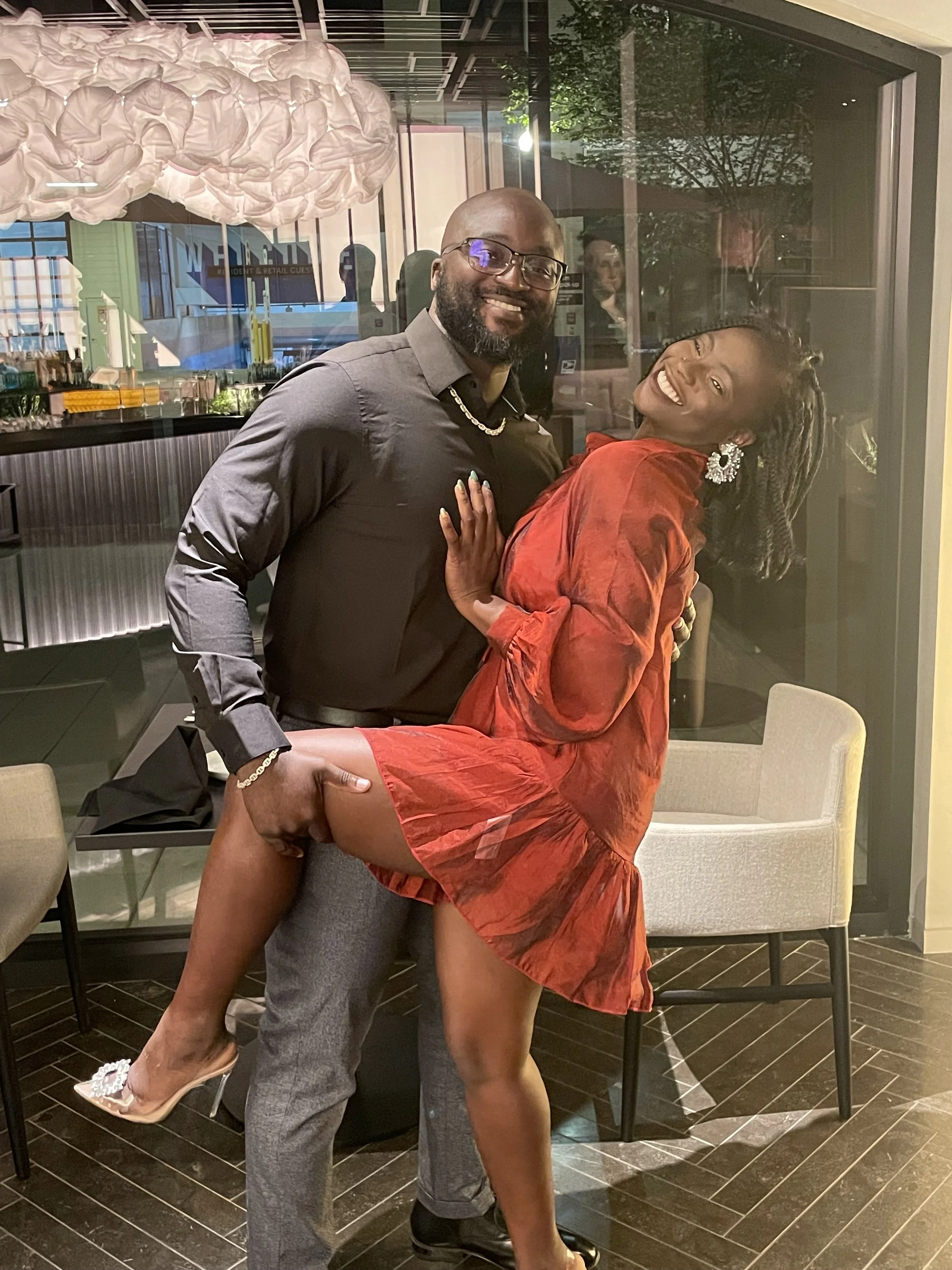  Liam is holding Moyo in his arms inside a modern restaurant with large glass windows and dark wooden flooring. 