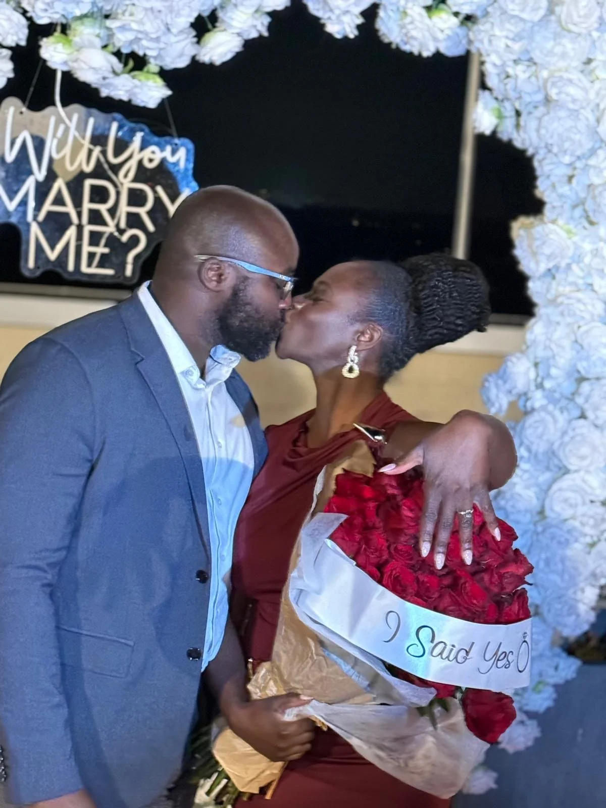 Liam and Moyo kissing at a wedding celebration, with the woman holding a bouquet of red roses and a sash reading 'I Said Yes'.
