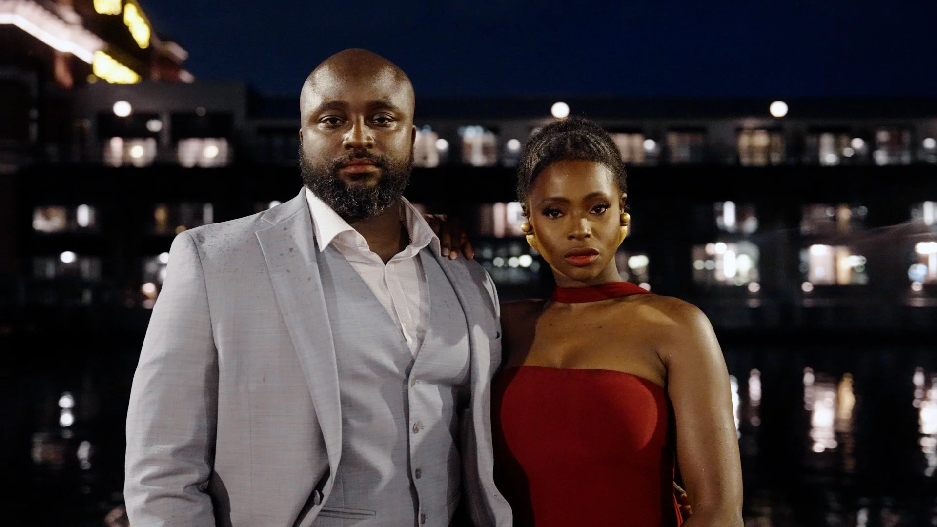 Liam in a light gray suit and Moyo in a strapless red dress stand close together outdoors at night in front of a dark background with lit windows reflecting on water.
