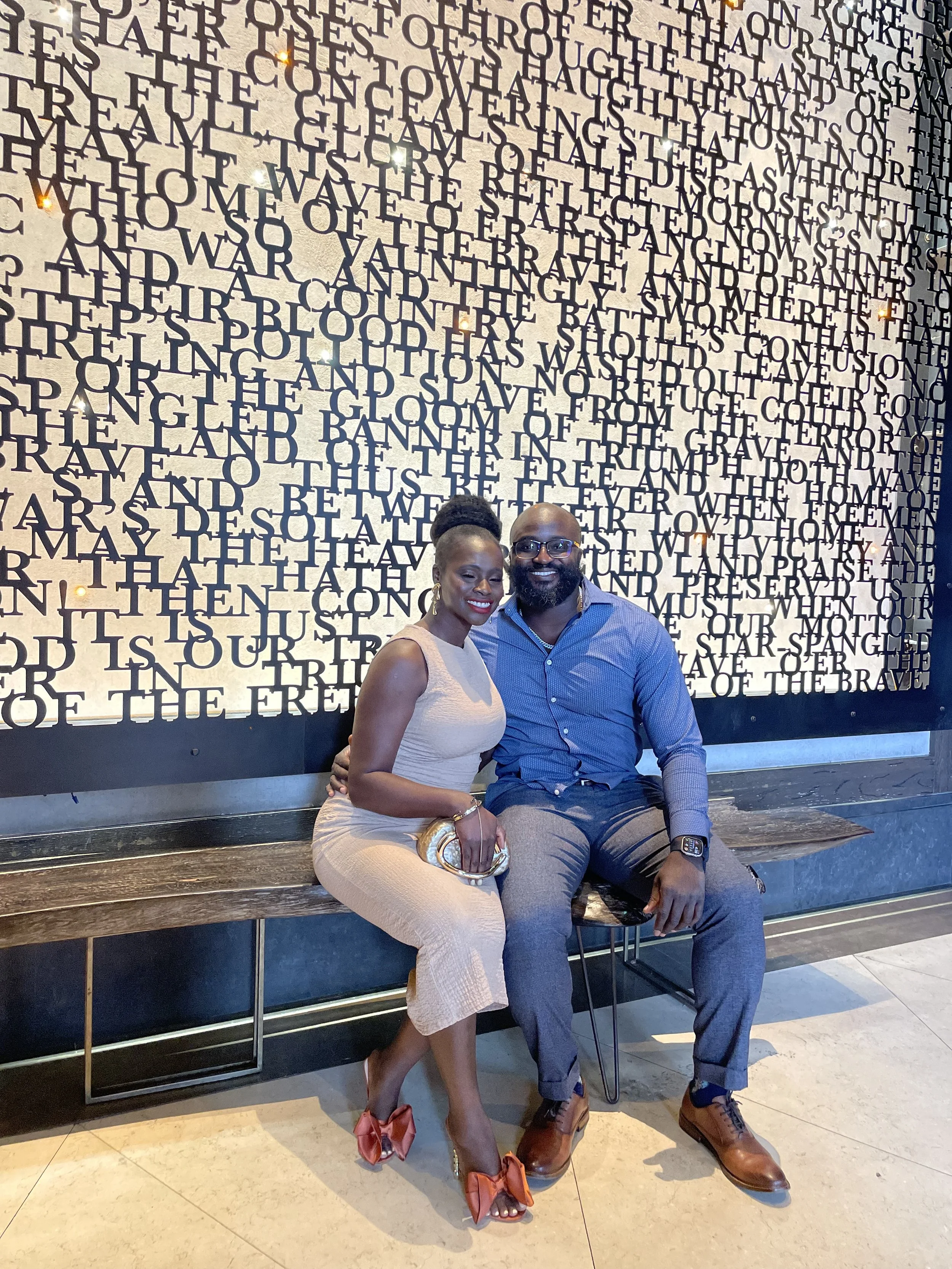 Liam and Moyo are sitting on a wooden bench in front of a wall with black text. They are smiling and posing together.
