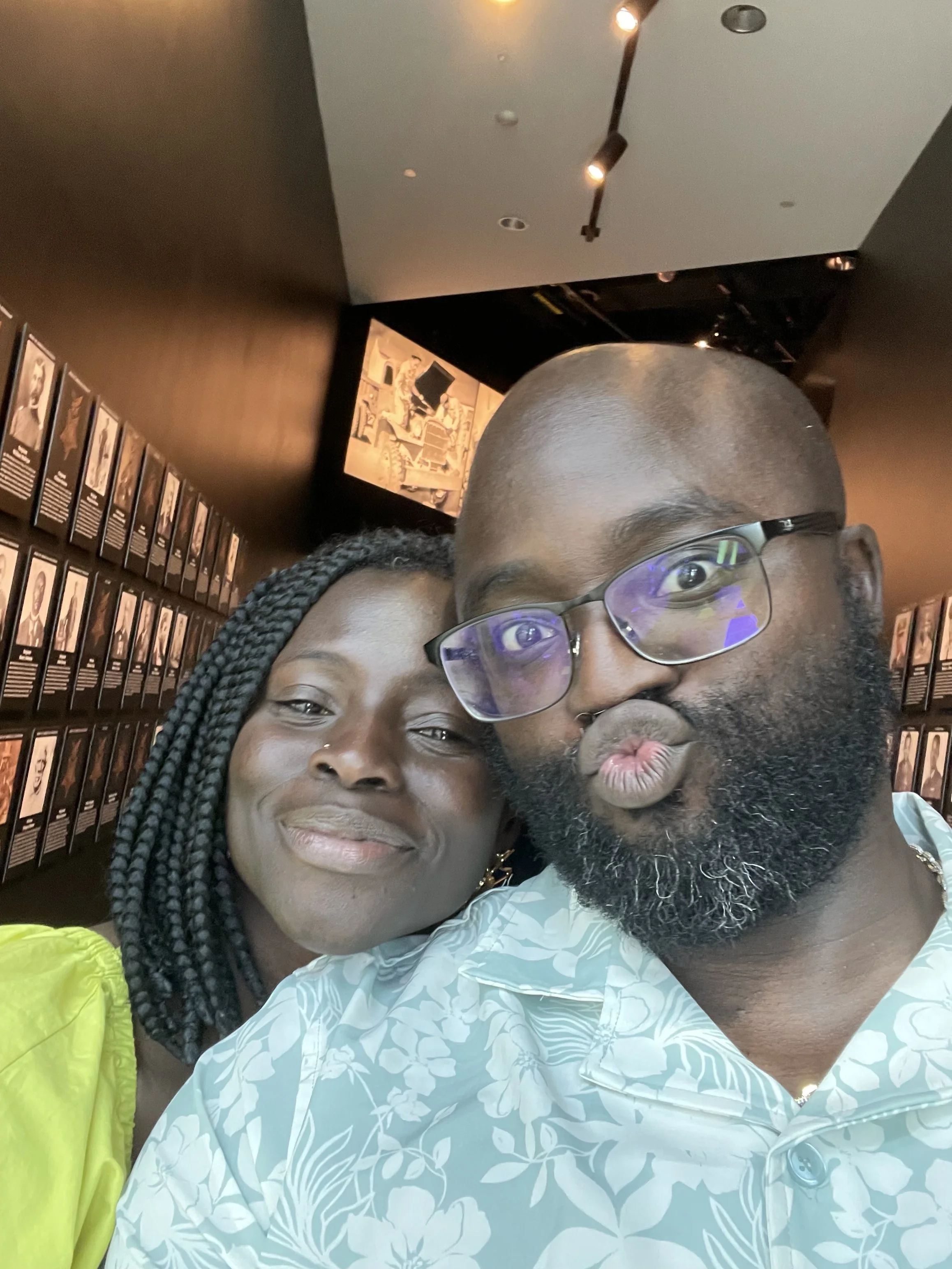 A close-up selfie of a smiling Moyo with braided hair and dark skin, and Liam with a beard, glasses, and dark skin, making a kissy face.