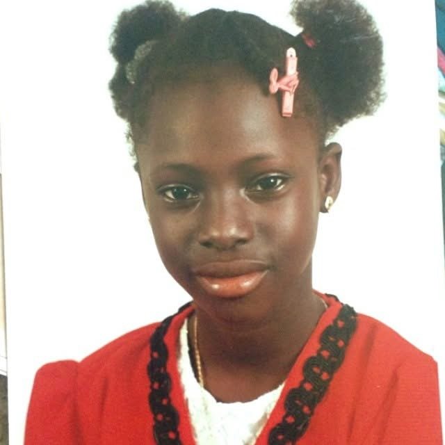 Young Moyo with dark hair styled in two puffy buns with pink hair clips, wearing a red garment with black trim, and a white top underneath.