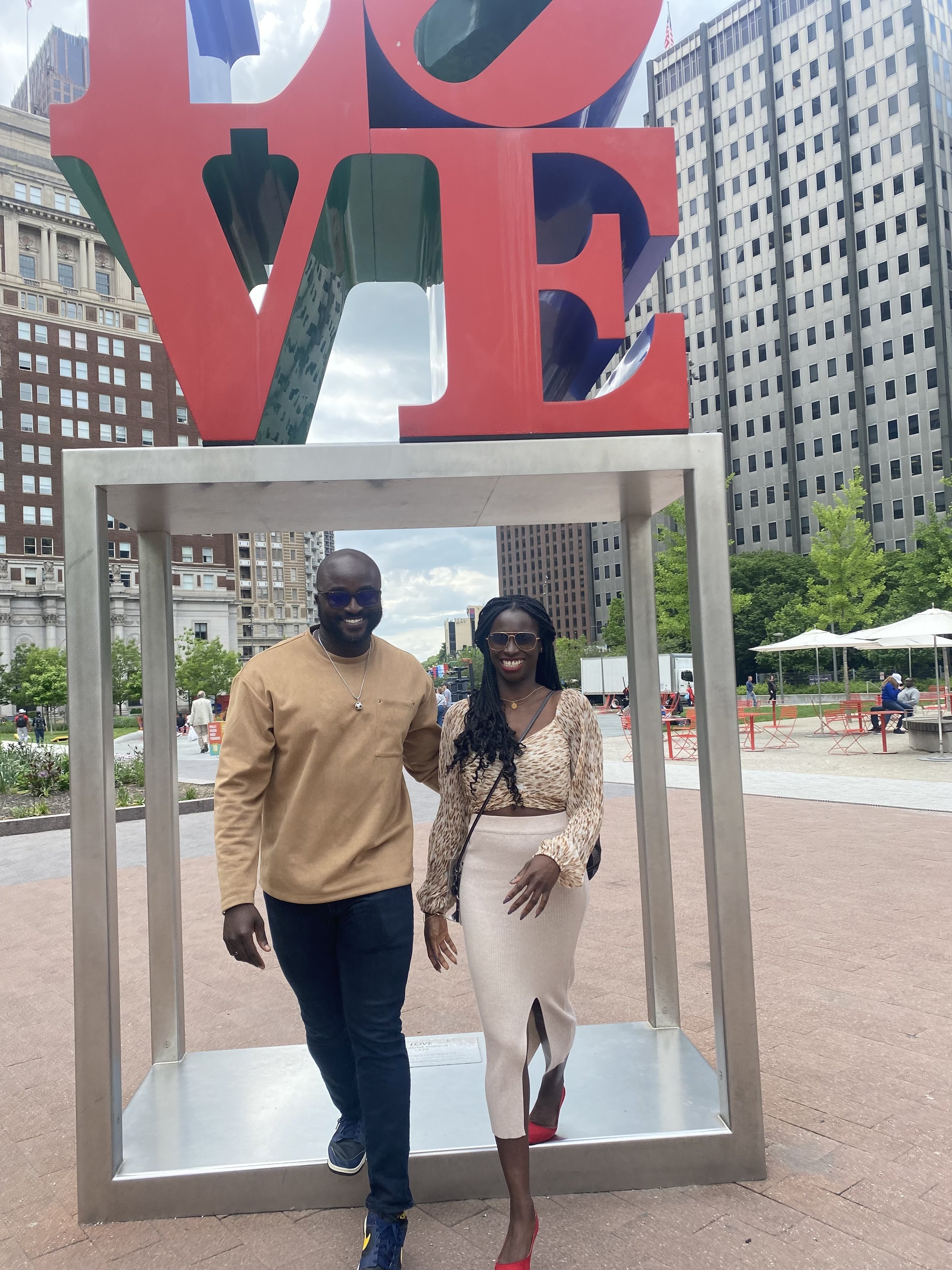 Moyo and Liam, smiling and walking together in front of a large sculpture of the word 'LOVE' in a city park. 