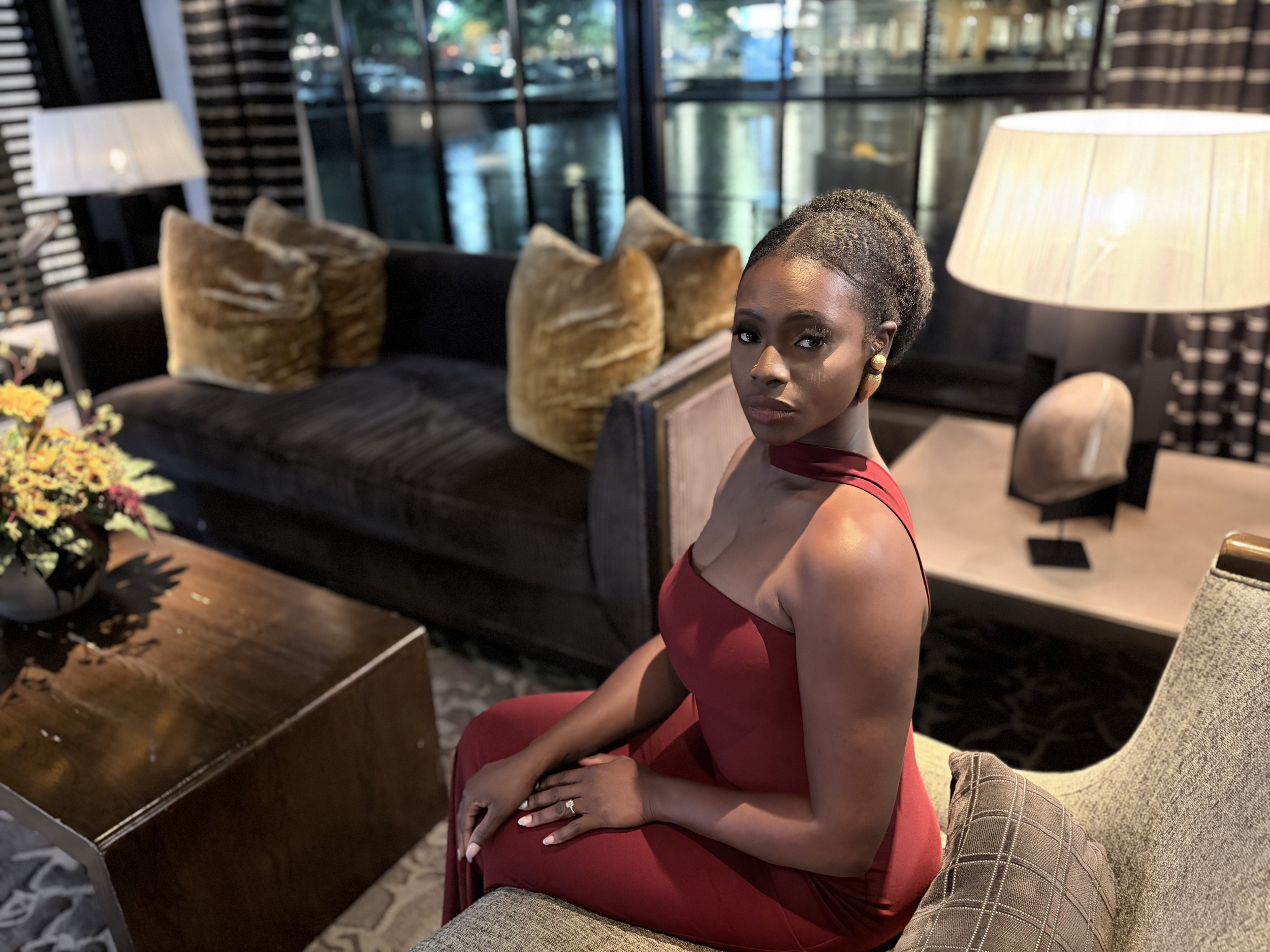 Moyo in a red dress sitting on a chair in a stylish living room with a sofa, large window, and warm lighting.