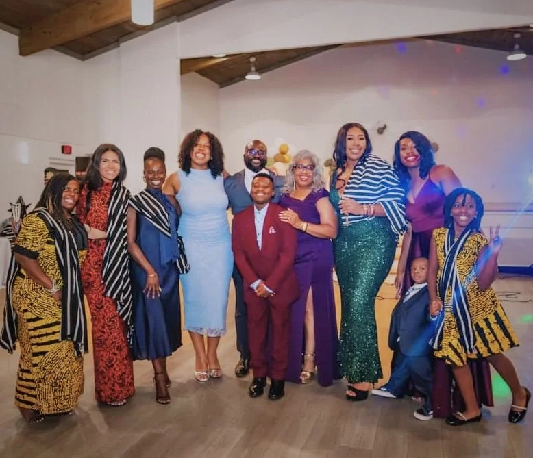 Moyo and Liam dressed in colorful formal attire at an indoor celebration or event with family.