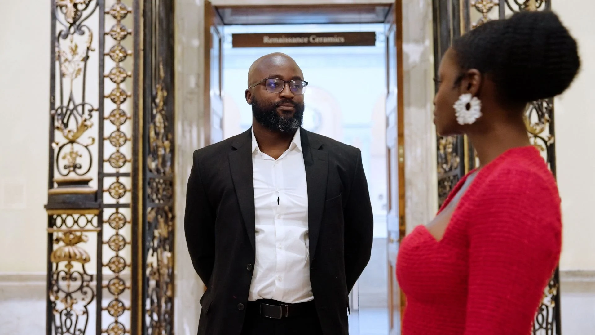 Liam and Moyo stand facing each other in an ornate indoor setting, possibly a museum or historical building, with a sign that reads 'Renaissance Ceramics' in the background.