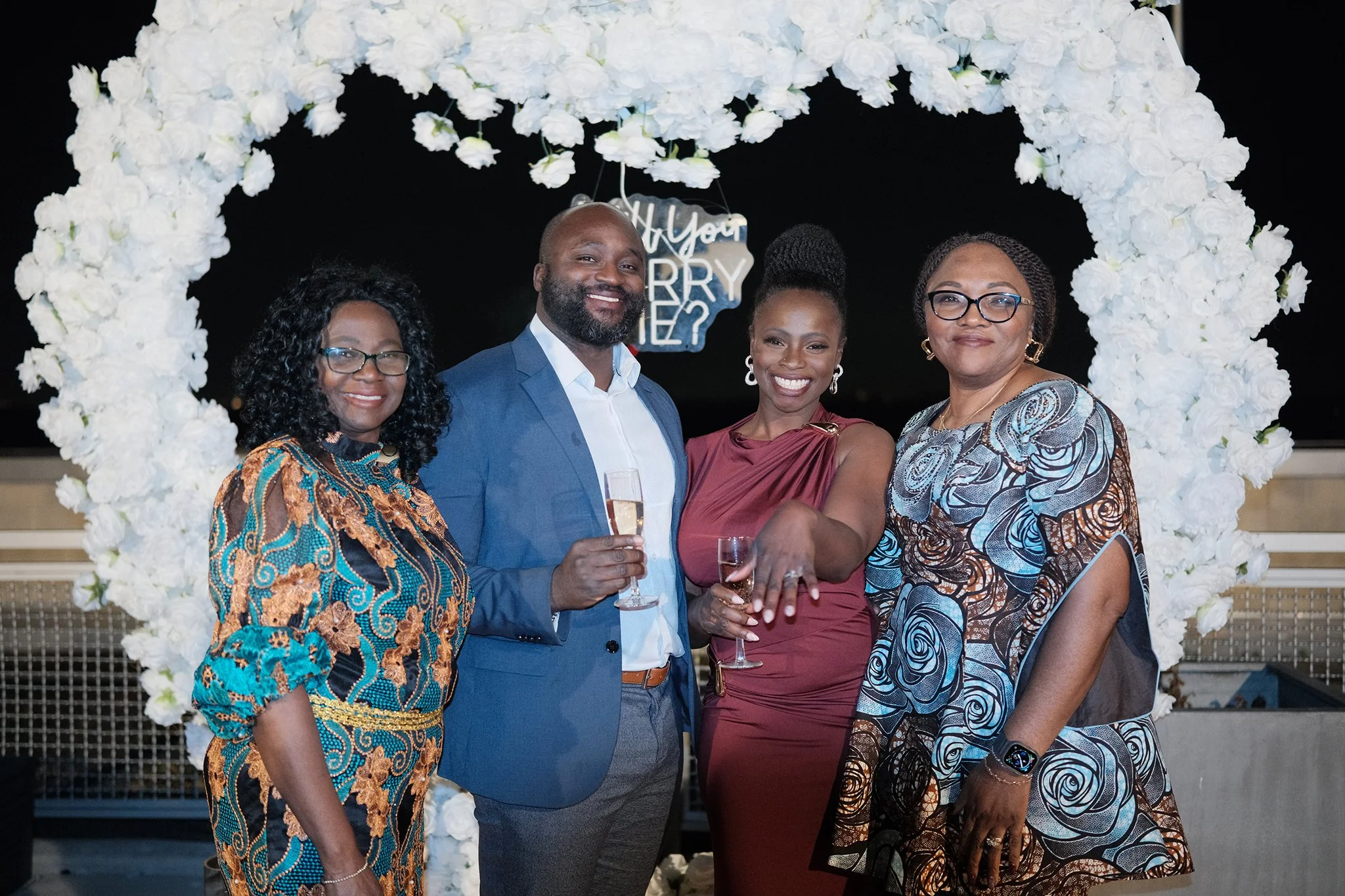 Moyo and Liam at their engagement celebration with their moms, standing under a white floral arch, holding glasses of champagne, with a sign that says 'Will You Marry?' in the background.