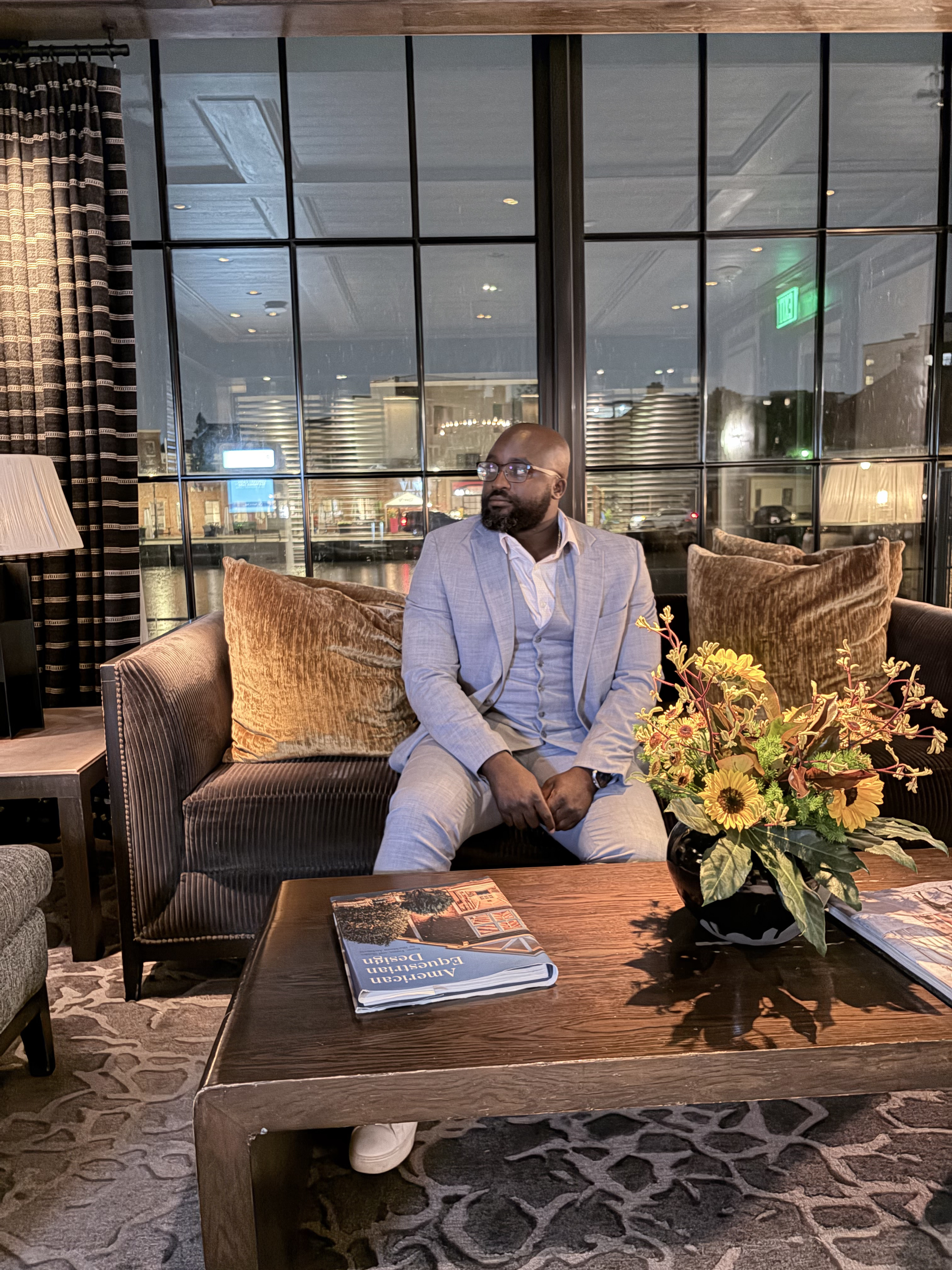 Liam, wearing a light gray blazer and white shirt, is seated on a brown couch inside a restaurant or lounge, with large windows behind him showing an urban cityscape at night, and a flower arrangement on a dark wooden coffee table in the foreground.